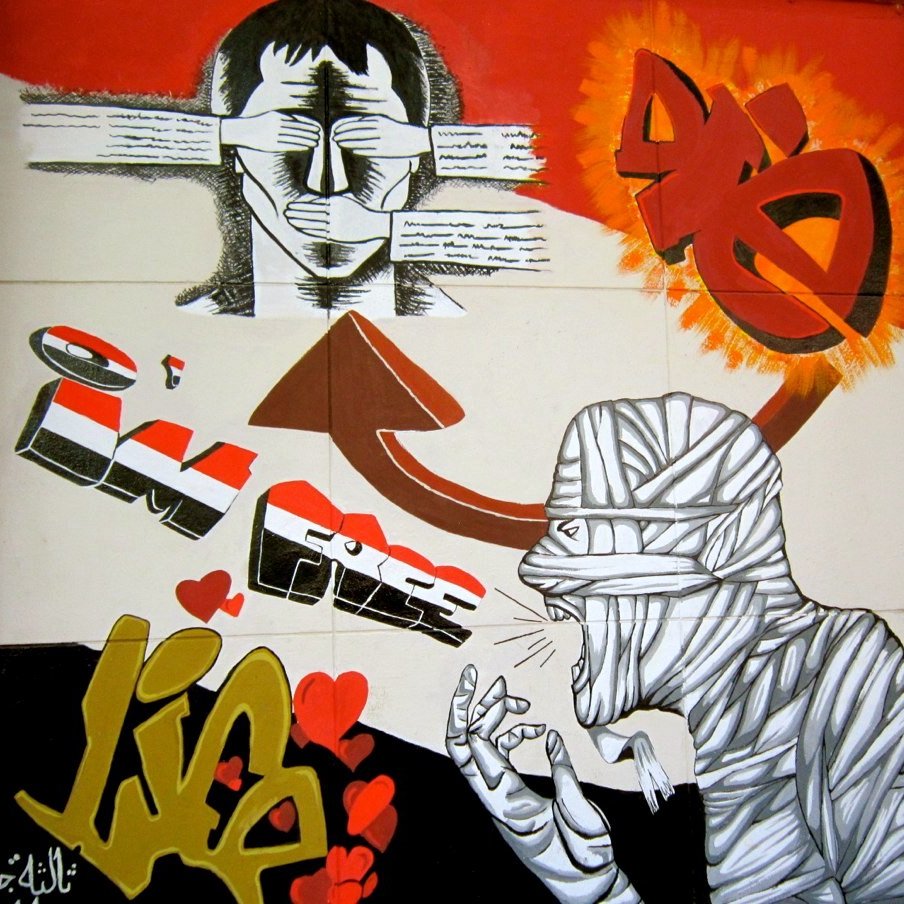 L'image représente une œuvre de street art colorée. On y voit un personnage avec des lunettes de soleil et une expression déterminée, entouré de textes. Au-dessus, une flèche pointe vers le haut avec l'inscription "I'M FREE" (je suis libre) en lettres stylisées. À droite, un autre personnage est représenté, désigné comme étant bandé, avec des gestes qui semblent exprimer une forte émotion. Le fond combine des couleurs vives comme le rouge et le jaune, avec des cœurs dessinés au bas, ce qui ajoute une touche plus douce à l'ensemble. L'œuvre montre un contraste entre la liberté et une certaine souffrance, évoquant des thèmes profonds.