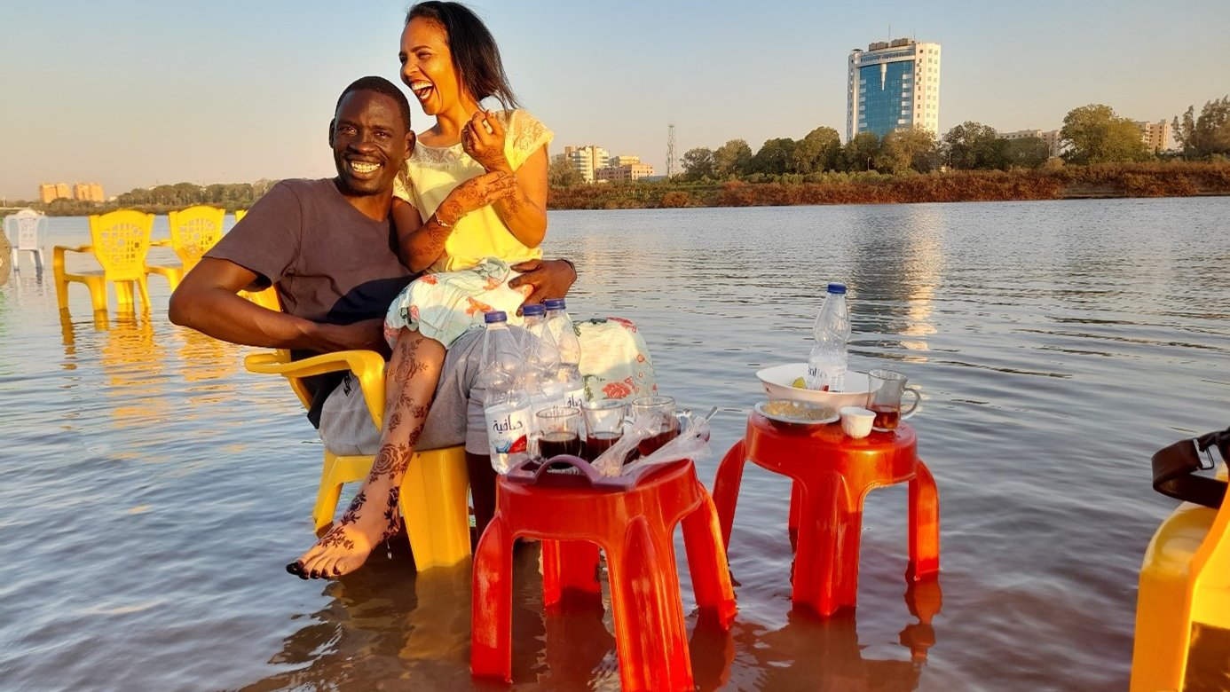L'image montre un couple assis sur des chaises jaunes au bord d'une étendue d'eau, probablement un lac ou une rivière. Ils semblent apprécier un moment convivial ensemble, riant et partageant des instants joyeux. À côté d'eux, il y a des tables avec des bouteilles d'eau et des verres. L'arrière-plan présente un paysage urbain avec des bâtiments, et la lumière du soleil crée une atmosphère chaleureuse et accueillante.