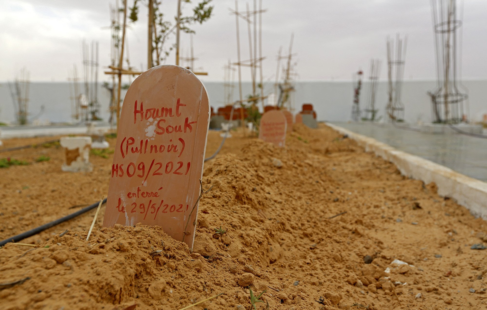 L'image montre un cimetière avec des tombes marquées par des plaques en terre cuite. Le sol est sablonneux et légèrement irrégulier, avec quelques plantes éparses. En arrière-plan, on remarque des structures en construction, probablement des bâtiments ou des monuments. Le ciel semble nuageux, créant une atmosphère calme mais mélancolique. Les inscriptions sur les plaques indiquent les noms et des dates, suggérant que ces êtres chers ont été récemment enterrés.