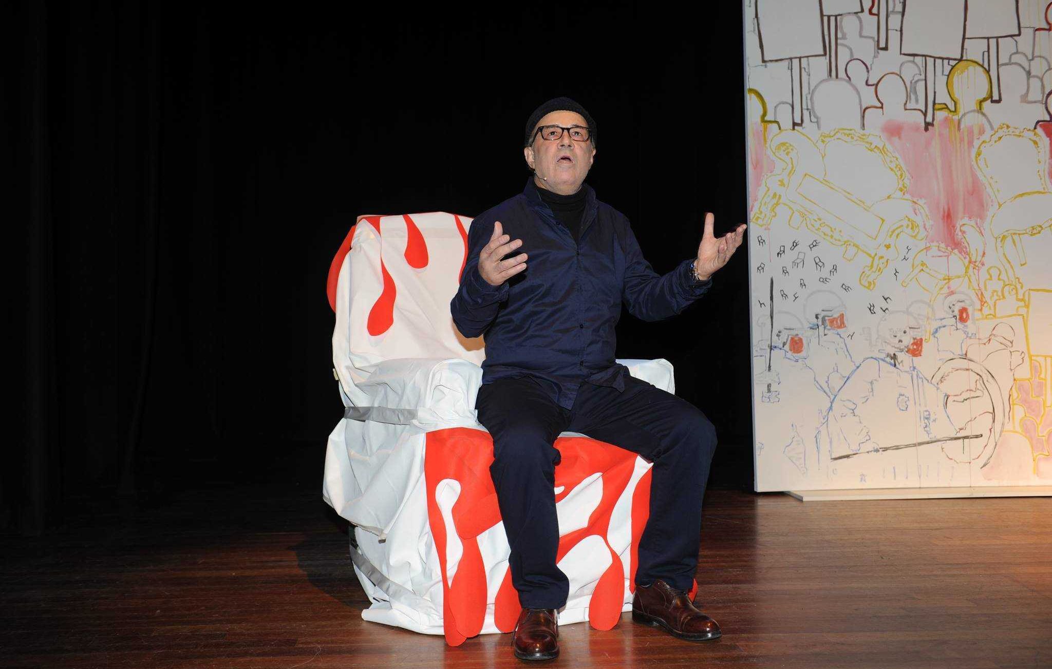 L'image montre un homme assis sur une chaise décorée de motifs rouges et blancs. Il est vêtu de vêtements sombres et porte un bonnet. L'homme a l'air expressif, gesticulant avec les mains, peut-être en train de parler ou de performer. En arrière-plan, on voit un panneau avec des dessins colorés et des formes abstraites. L'atmosphère semble être celle d'une scène de théâtre.