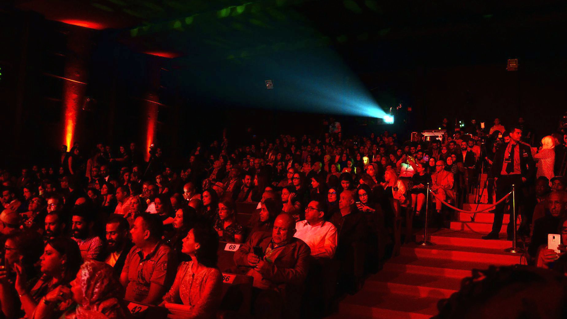L'image montre un auditorium rempli de spectateurs. La salle est faiblement éclairée avec des lumières rouges qui créent une ambiance chaleureuse et dynamique. Au centre, un projecteur émet un faisceau lumineux, ajoutant à l'atmosphère du moment. Les spectateurs, composés d'hommes et de femmes, semblent attentifs et engagés, certains tenant des téléphones. L'escaliers sur le côté de la scène permet l'accès à d'autres niveaux. L'ensemble de la scène évoque un événement artistique ou culturel.