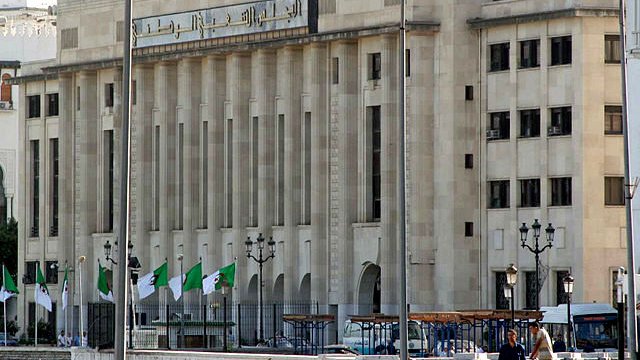 L'image montre un bâtiment majestueux avec une architecture moderne. La façade est claire, avec de grandes fenêtres et des lignes géométriques. On peut également voir des drapeaux algériens flottant devant le bâtiment. Il y a des lampadaires et une ambiance urbaine. Ce contraste entre le style architectural et le décor environnant suggère un lieu important, probablement lié aux institutions gouvernementales.