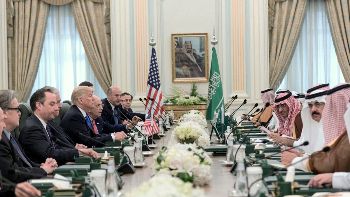 در این تصویر، یک جلسه رسمی در یک اتاق بزرگ برگزار میشود. در سمت چپ، تعدادی از مقامهای آمریکایی نشستهاند و پرچم ایالات متحده در کنار آنها قرار دارد. در سمت راست، مقامهای سعودی نشستهاند و پرچم عربستان سعودی به چشم میخورد. بر روی میز، گلهای سفید و دیگر وسایل اداری وجود دارد. این جلسه نشاندهنده تعاملات دیپلماتیک و همکاریهای بینالمللی بین دو کشور است. دیوارهای اتاق با پردههای زیبا تزئین شده و تصویری بزرگ از یک شخصیت تاریخی در پسزمینه قرار دارد.