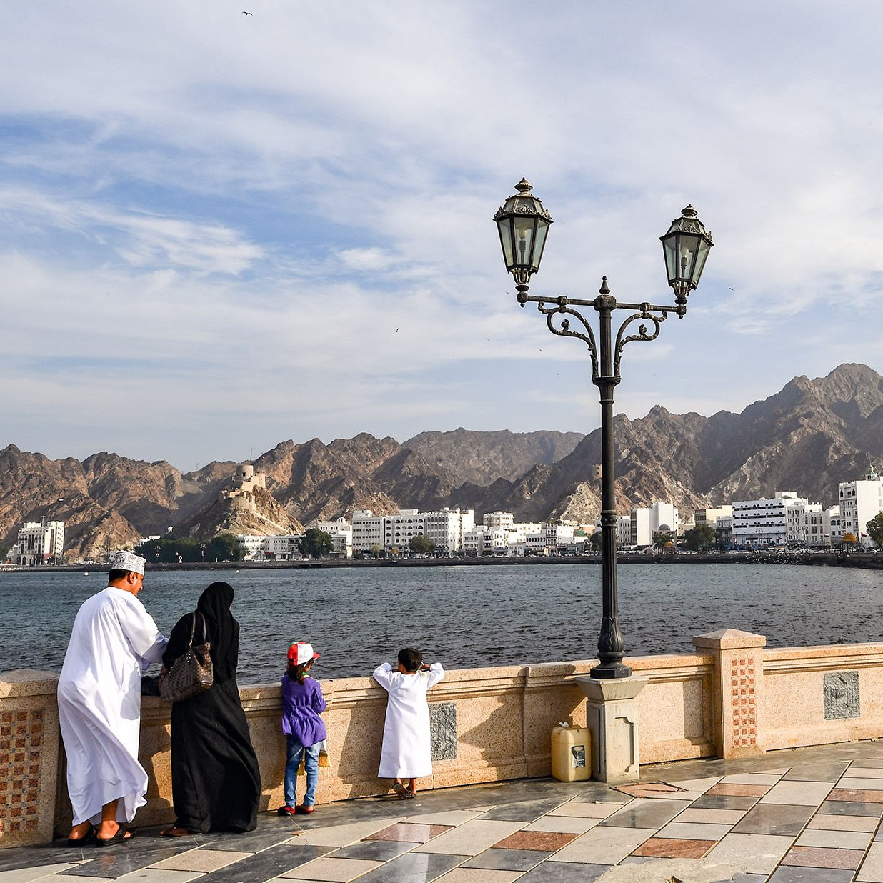 تظهر الصورة منظرًا جميلًا لواجهة بحرية في منطقة جبلية. في المقدمة، يقف شخصان وطفلان على حافة المياه، حيث يستمتع الجميع بالمنظر. الشخصان يرتديان ملابس تقليدية، بينما يرتدي الأطفال ملابس ملونة. في الخلفية، تظهر الجبال الشاهقة ومناظر المدينة مع بعض المباني البيضاء. السماء زرقاء مع غيوم خفيفة، مما يعطي الصورة شعورًا بالهدوء والطبيعة الجميلة. عمود إنارة يضيف لمسة جمالية للمشهد.