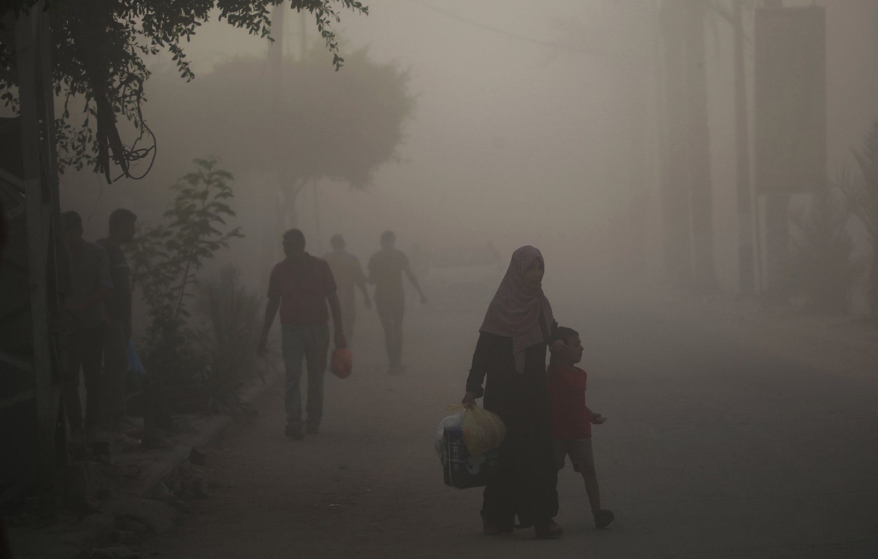 L'immagine mostra una scena di una strada avvolta nella nebbia o nella polvere. In primo piano, una donna con un velo scuro tiene la mano di un bambino, mentre camminano insieme. Sullo sfondo, si possono vedere altre persone che camminano, ma sono poco visibili a causa della foschia. Gli alberi e alcune piante ai lati della strada creano un'atmosfera un po' opprimente. La luce sembra attenuata, dando un'impressione di desolazione.