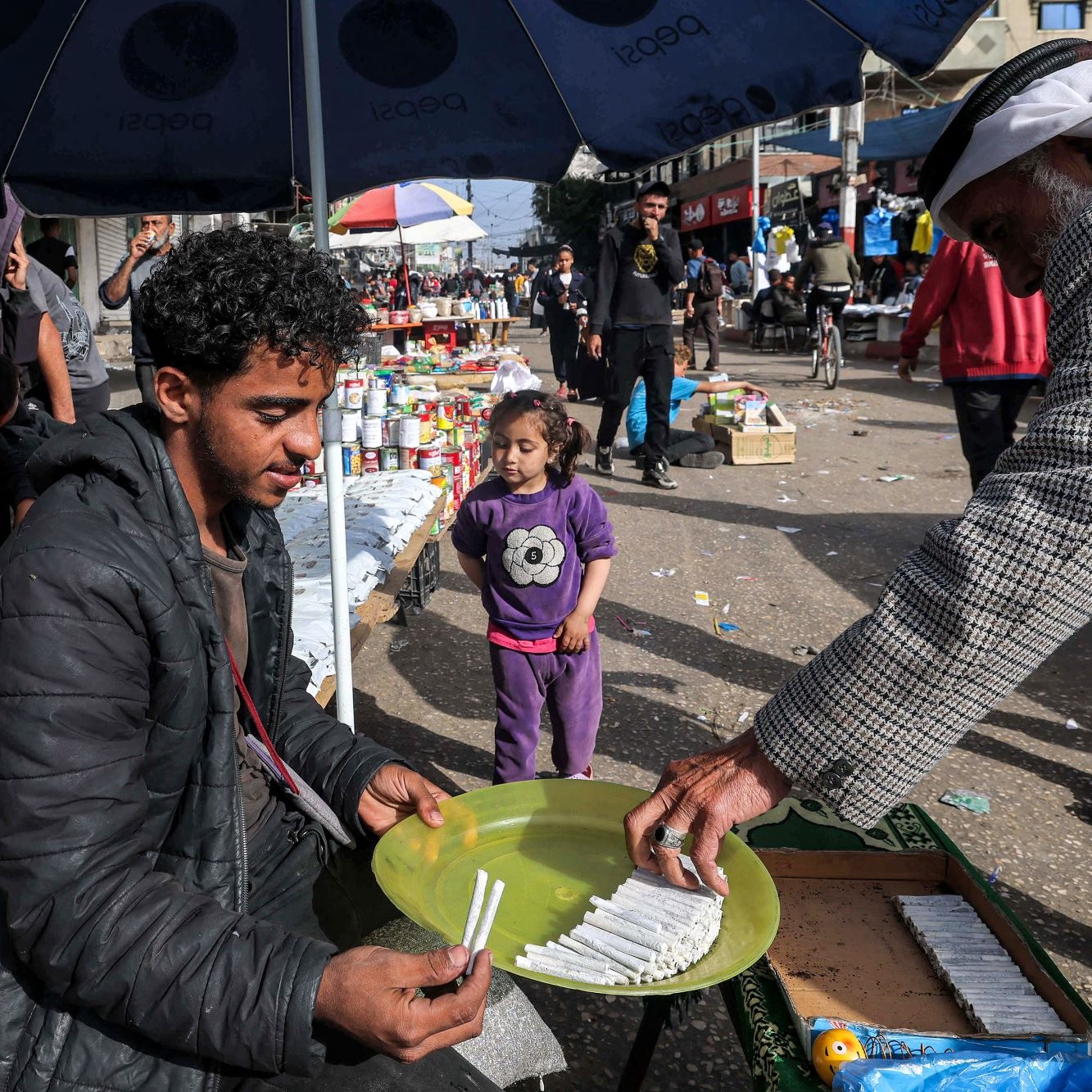 L'immagine mostra una scena di mercato all'aperto, probabilmente in una città. Un giovane uomo è seduto su un contenitore di legno, di fronte a un tavolo, mentre tiene in mano un piatto. Sta vendendo o mostrando dei prodotti, probabilmente cibo o articoli vari. Accanto a lui, un uomo anziano, vestito con indumenti tradizionali, si avvicina per prendere qualcosa dal piatto. Sullo sfondo, ci sono altre persone che passeggiano e alcune bancarelle con vari articoli in esposizione. Una bambina, vestita con una maglietta, osserva curiosa la scena. La luce del giorno illumina vividamente l'ambiente, creando un'atmosfera vivace e animata.