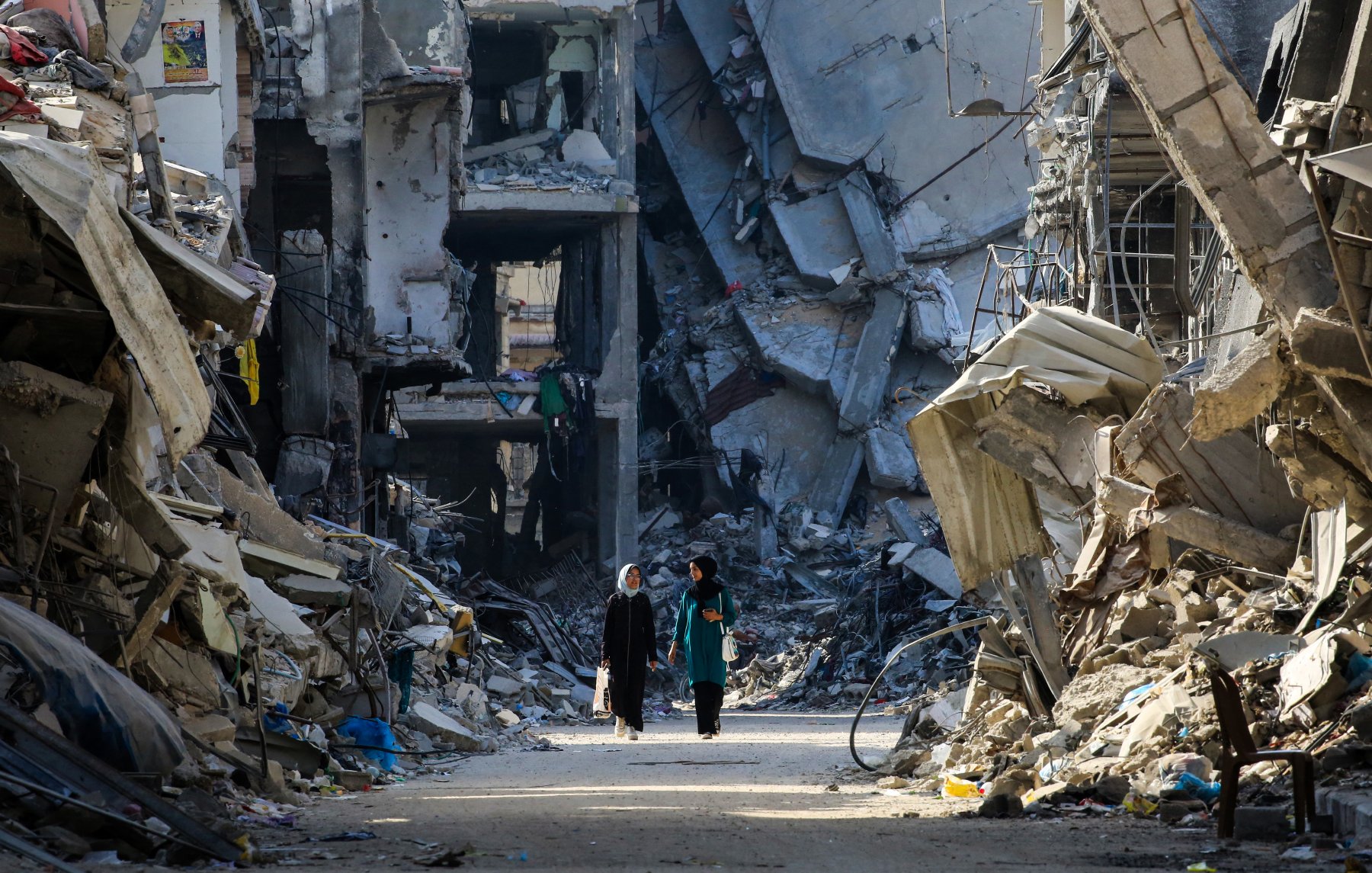 L'immagine mostra due persone che camminano in una strada distrutta da un conflitto, circondate da edifici in rovina. Le strutture sono parzialmente crollate e si vedono macerie sparse ovunque, creando un'atmosfera di desolazione e devastazione. La scena evidenzia la gravità della situazione, con un forte contrasto tra le persone che camminano e l'ambiente circostante, che riflette la difficoltà e la sofferenza di un'area colpita dalla guerra.