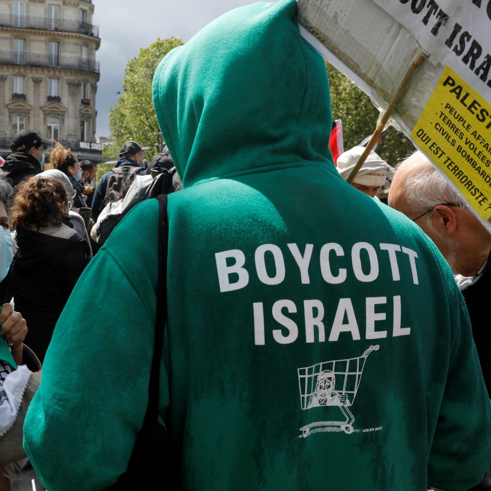 The image depicts a gathering or protest where individuals are expressing their political views. In the foreground, a person wearing a green hoodie that reads "BOYCOTT ISRAEL" is facing another individual. The latter is wearing a face mask and has a traditional black-and-white patterned scarf. The setting appears to be in a public space, with banners and flags present in the background, indicating a demonstration related to Palestinian rights or opposition to certain policies regarding Israel.