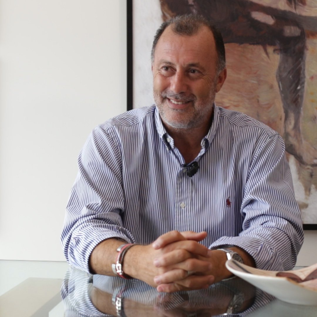 The image shows a man sitting at a glass table with a friendly smile. He is wearing a blue-striped shirt and has short, dark hair. Behind him, there are two framed artworks on the wall, one of which appears to be a sketch or abstract piece. The setting looks modern and well-lit, suggesting a comfortable and professional atmosphere. The man seems relaxed and engaged in conversation.