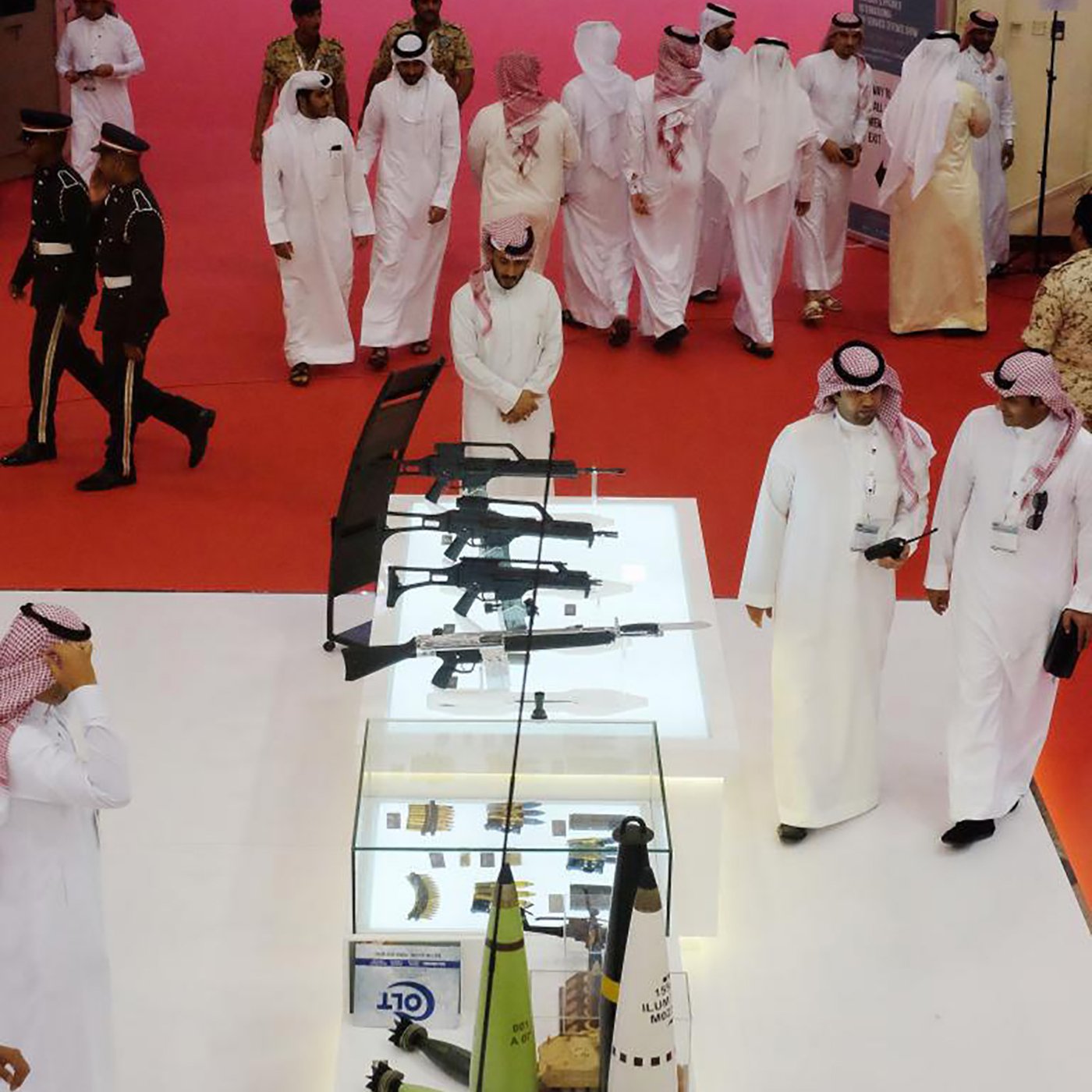 L'image montre un rassemblement où plusieurs personnes, probablement en tenue traditionnelle, se déplacent dans un espace décoré. Au centre, il y a une vitrine en verre contenant diverses armes, mettant en avant des fusils et d'autres équipements. L'ambiance semble formelle et le sol est recouvert d'un tapis rouge. Des membres de la sécurité ou des uniformes militaires peuvent être aperçus en arrière-plan, soulignant l'importance de l'événement.