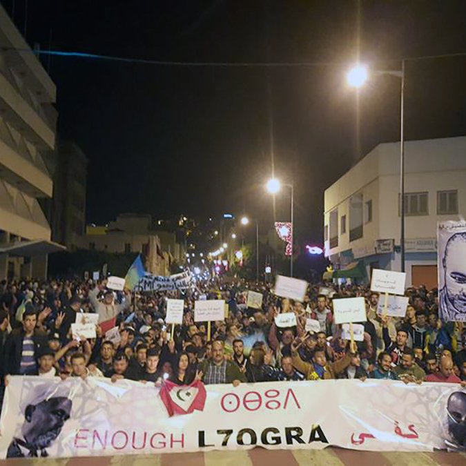 L'image montre une grande manifestation nocturne dans une rue animée. Beaucoup de personnes sont rassemblées, tenant des pancartes et banderoles. On peut voir des portraits de plusieurs individus exposés parmi les manifestants. L'atmosphère semble énergique et engagée, avec des participants qui expriment leurs revendications. Des lumières artificielles illuminent la scène, créant une ambiance dynamique.