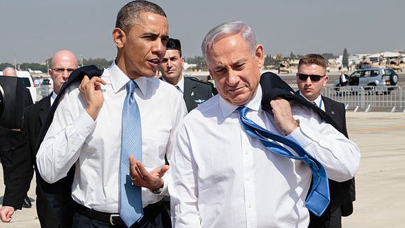 L'image montre deux hommes, habillés en costume, marchant ensemble sur un tarmac. L'un porte une cravate bleue et l'autre une cravate claire. Ils semblent engagés dans une conversation. En arrière-plan, on peut apercevoir des membres de la sécurité et des véhicules. Le ciel est clair, et l'environnement semble être un aéroport.