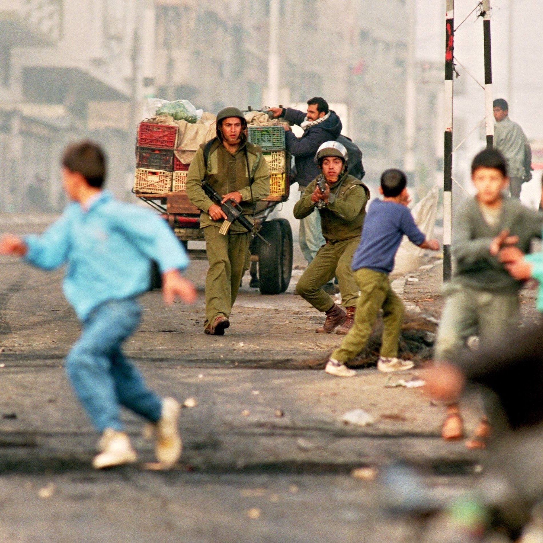L'immagine ritrae una scena di conflitto in un'area urbana. Due soldati in uniforme stanno correndo, uno dei quali sembra puntare un'arma. Dietro di loro, un carro con casse di frutta è visibile. In primo piano, dei bambini stanno fuggendo, mostrando un'espressione di paura. L'ambiente circostante appare caotico, con una luce che suggerisce un'atmosfera di tensione. La scena comunica chiaramente una situazione di crisi e instabilità.