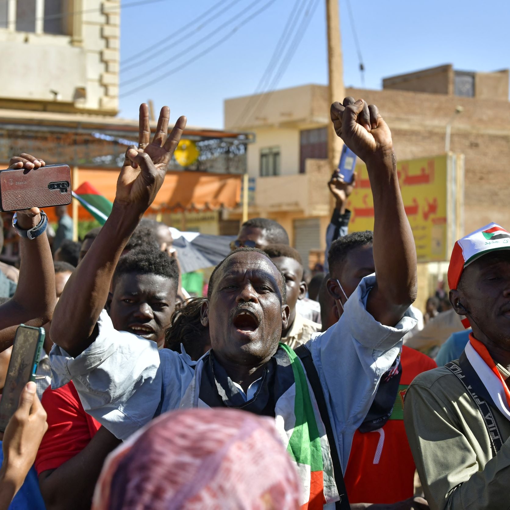 L'image montre une foule de manifestants lors d'une manifestation. Ils semblent exprimer leur colère ou leur détermination, avec des poings levés et des expressions intenses. Certaines personnes tiennent des téléphones portables, tandis qu'une autre utilise un mégaphone pour s'exprimer. En arrière-plan, on aperçoit des bâtiments et des drapeaux, ce qui donne une idée d'un contexte de protestation pour une cause déterminée. L'atmosphère semble énergique et passionnée.