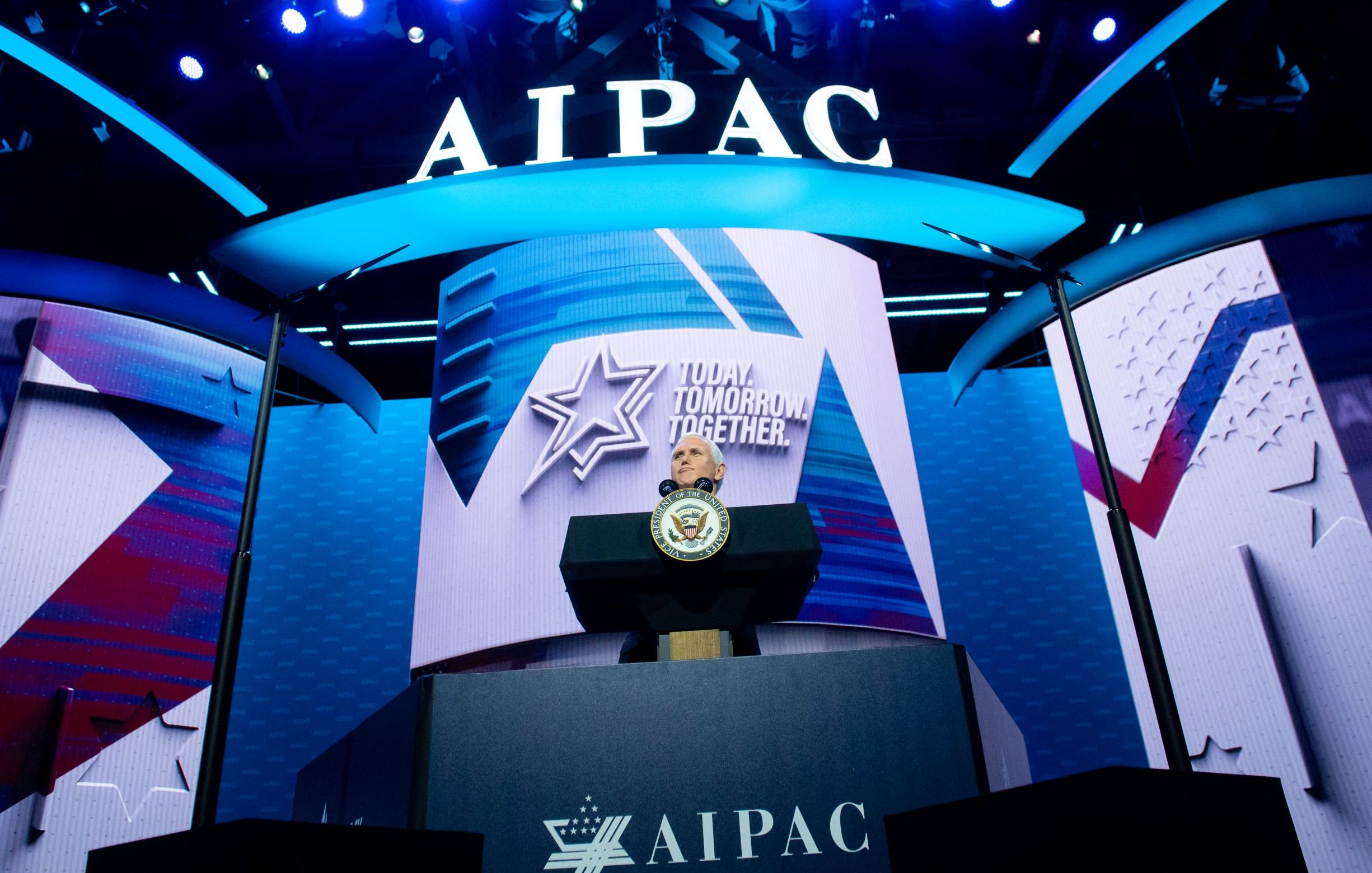 L'image montre une scène de conférence avec un podium bien en vue, sur lequel se trouve un sceau officiel. En arrière-plan, on peut voir un grand écran affichant le logo de l'AIPAC (American Israeli Public Affairs Committee), ainsi qu'un slogan qui pourrait être une thématique de l'événement. L'éclairage est dynamique, avec des lumières bleues qui créent une ambiance visuelle engageante. Cette configuration suggère qu'il s'agit d'un discours ou d'une présentation importante.