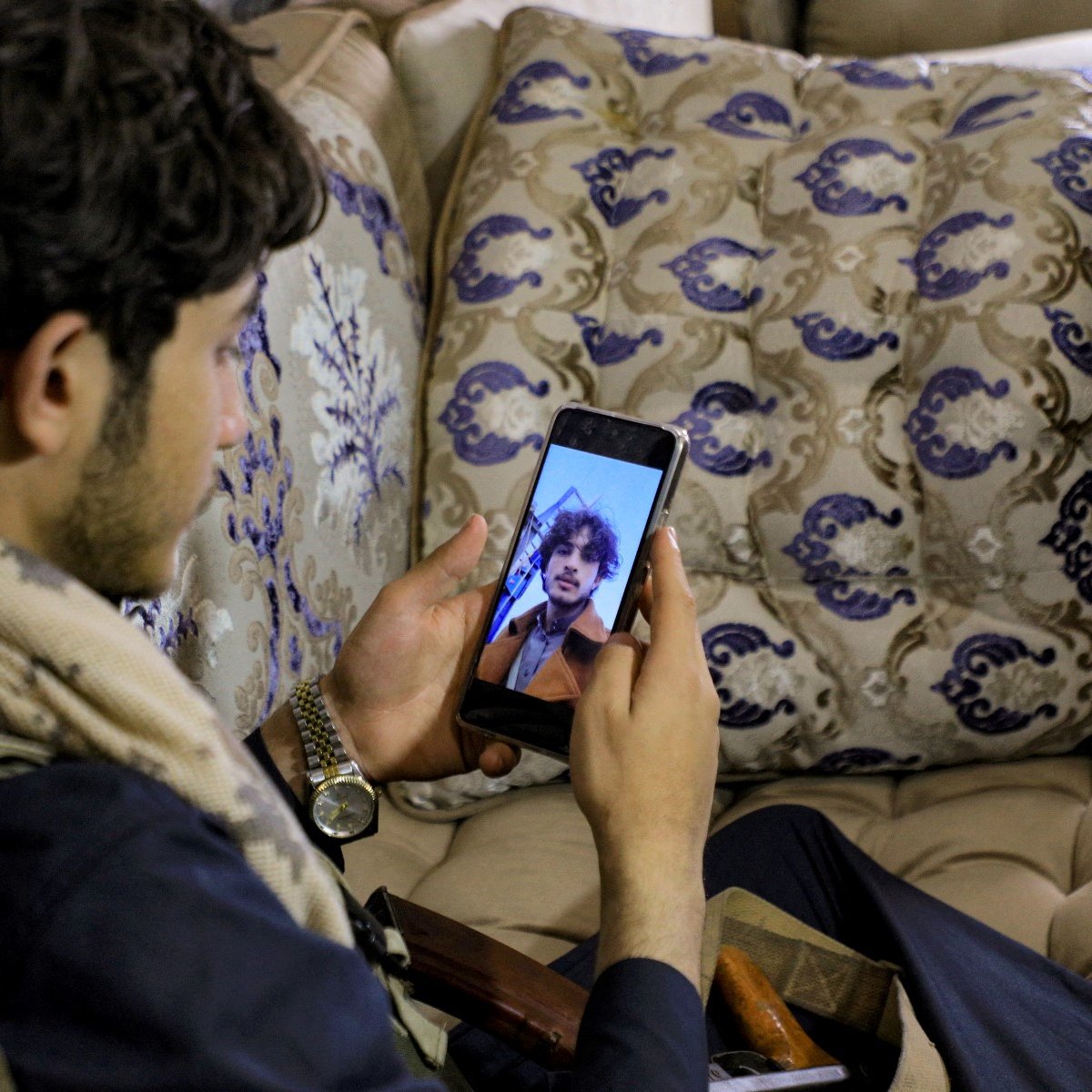 L'image montre un jeune homme assis sur un canapé décoré, regardant son téléphone portable. Il porte une écharpe et semble contemplatif. À côté de lui, on peut apercevoir une boîte et un objet, peut-être une arme ou un accessoire. Le décor est chaleureux et traditionnel, avec des coussins ornés de motifs élégants.