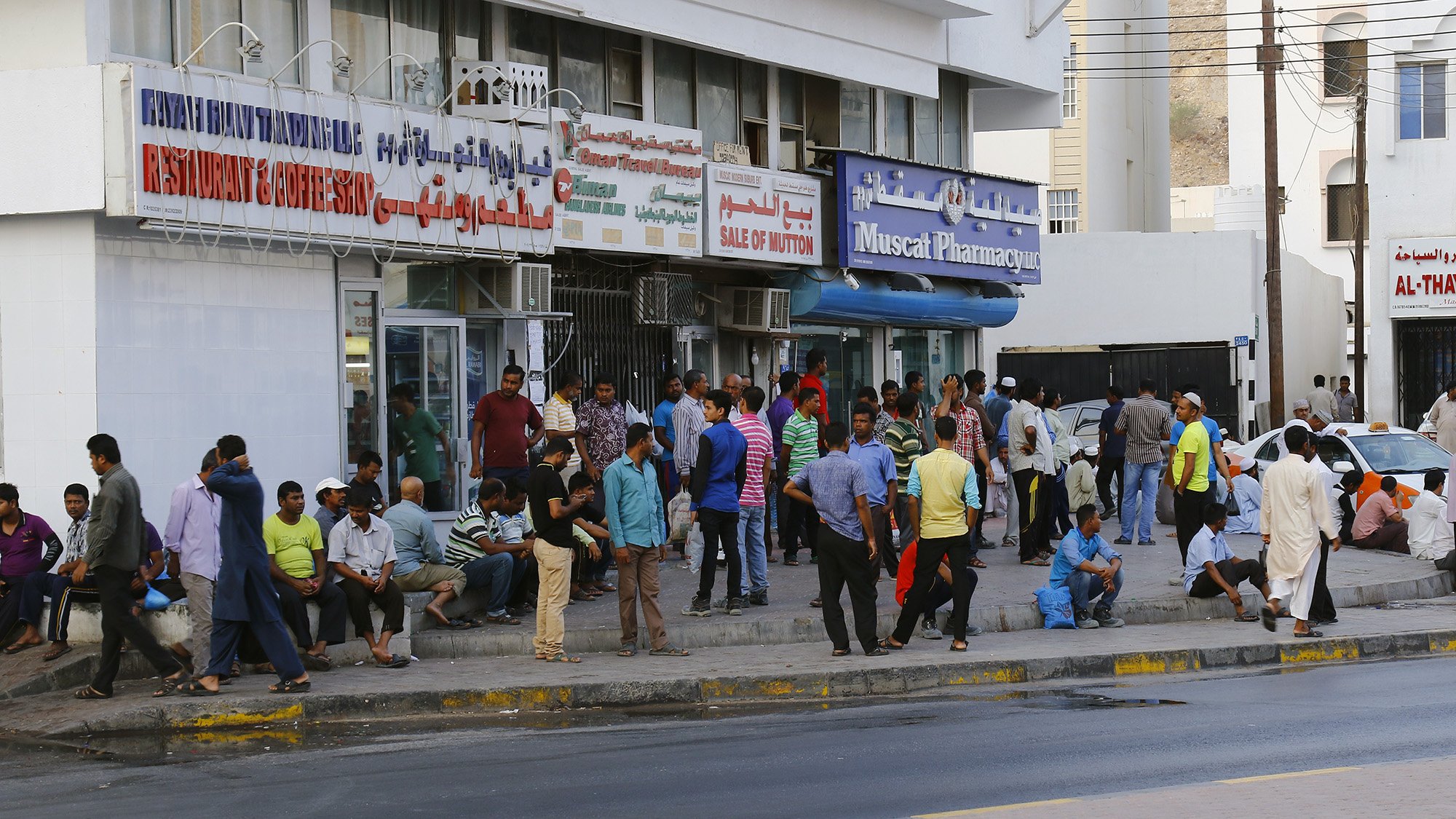تظهر الصورة مشهداً من شارع في منطقة حضارية مزدحمة. يوجد العديد من الأشخاص يتجمعون بالقرب من storefrontات ومتاجر متنوعة. يمكن رؤية مجموعة من الرجال والمارة يجلسون أو يقفون، وبعض الناس مشغولون بالتحدث مع بعضهم. يضم المشهد أيضاً لافتات باللغة العربية تشير إلى المتاجر والمرافق. الجو يبدو نشطًا، مع حركة مرور على الشارع في الخلفية.