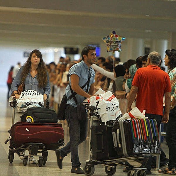 L'image montre une scène dans un aéroport, avec des voyageurs. À l'avant, un homme pousse une chariot de bagages et une femme derrière lui, également avec un chariot. Ils semblent attendent ou se déplacent parmi d'autres passagers. En arrière-plan, on aperçoit une longue file de personnes, probablement en attente de faire enregistrer leurs bagages ou de passer la sécurité. L'ambiance semble animée, typique des aéroports.