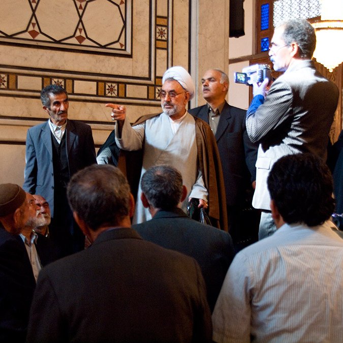 تظهر الصورة مجموعة من الأشخاص في مكان داخلي، حيث يركز المشهد على رجل يرتدي لباسًا تقليديًا يشير بيده ويتحدث إلى الحضور. يبدو هناك تفاعل قوي بين المتحدث والجمهور، الذين يظهرون جالسين أو واقفين في صفوف أمامه. في الخلفية، يمكن رؤية بعض الأشخاص الآخرين، وبعضهم يرتدي ملابس تقليدية. الأجواء تعكس طابعًا ثقافيًا أو دينيًا، مع تفاصيل معمارية تزين الجدران.