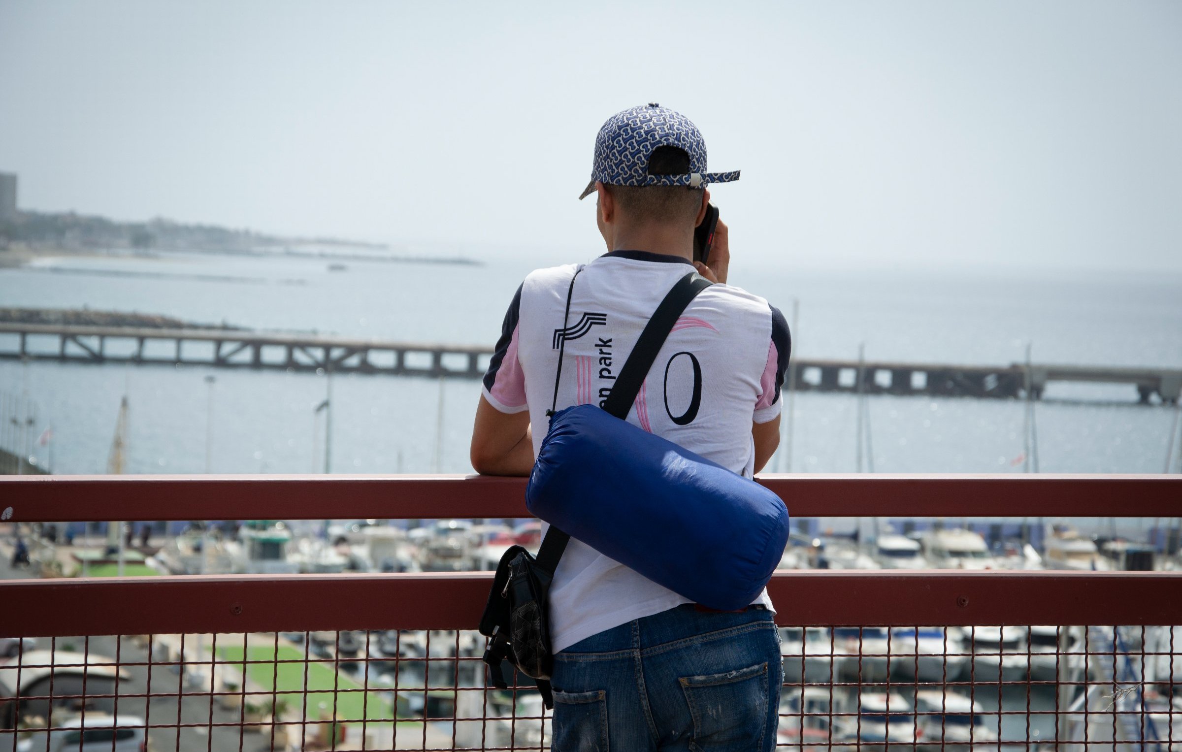 Un homme regarde la mer depuis un pont, avec un port et des bateaux en arrière-plan.