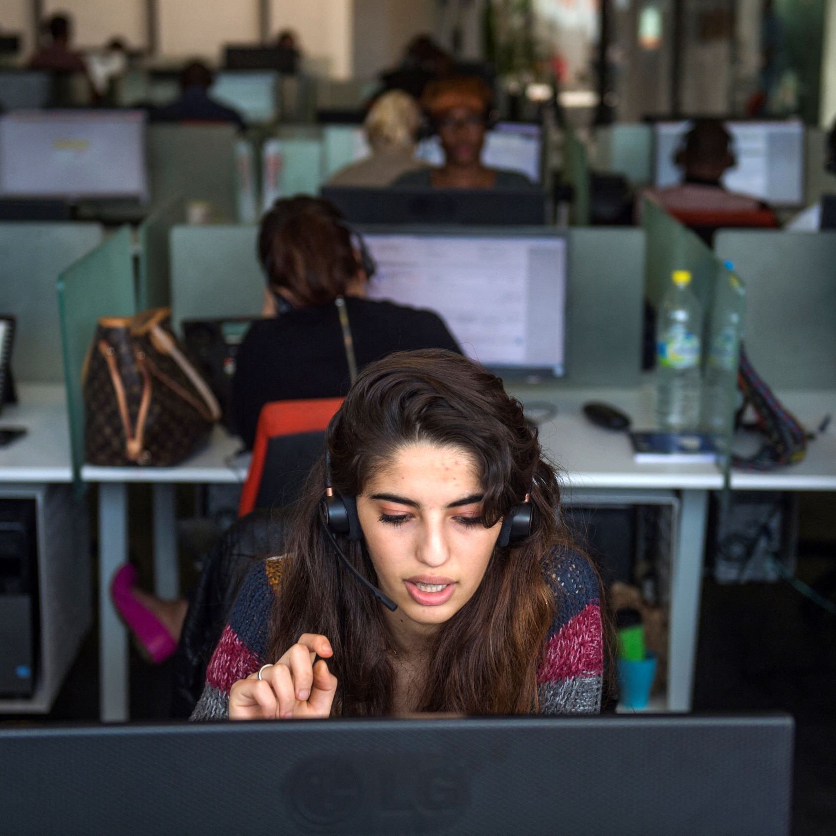 تظهر الصورة مكتبا حديثا في بيئة عمل، حيث توجد مجموعة من الأشخاص يعملون في مراكز اتصال. في المقدمة، تجلس شابة ترتدي سماعة رأس وتتحدث إلى ميكروفون، بينما تركز على شاشة الكمبيوتر أمامها. خلفها، يمكن رؤية زملاء آخرين يعملون، كلٌ منهم متواجد أمام حاسوبه. يظهر في الخلفية بعض الأغراض الشخصية مثل الحقائب، مما يضيف طابعاً إنسانياً للمكان. الإضاءة في المكتب طبيعية وتساهم في خلق جو عملي.