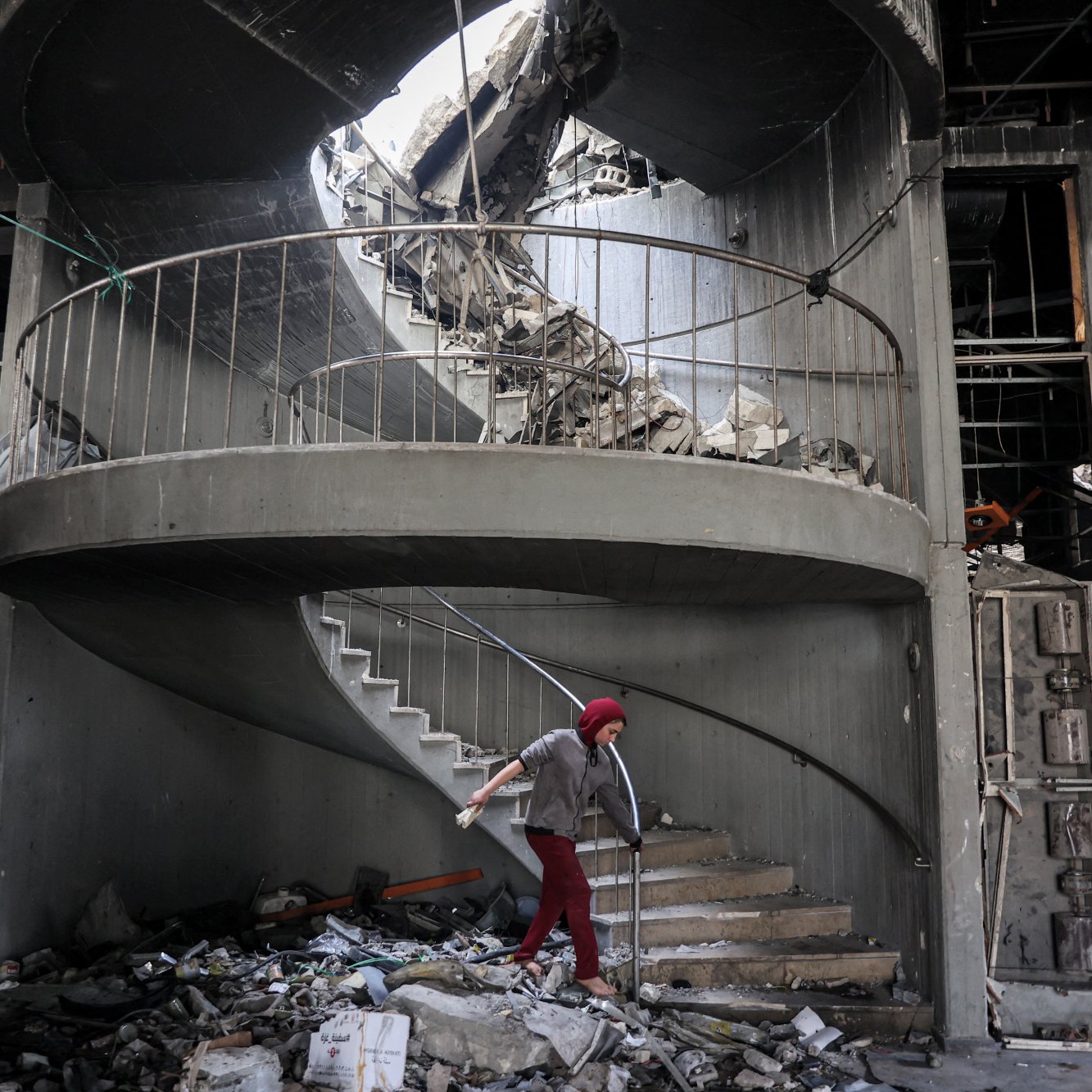 L'image montre un escalier en colimaçon partiellement détruit, avec des débris éparpillés autour. Le plafond a subi d'importants dégâts, laissant apparaître un trou par lequel la lumière pénètre. Un individu, utilisant des béquilles, se déplace prudemment sur les marches. L'ambiance générale évoque des conditions de délabrement et de désolation, probablement à la suite d'un événement catastrophique.