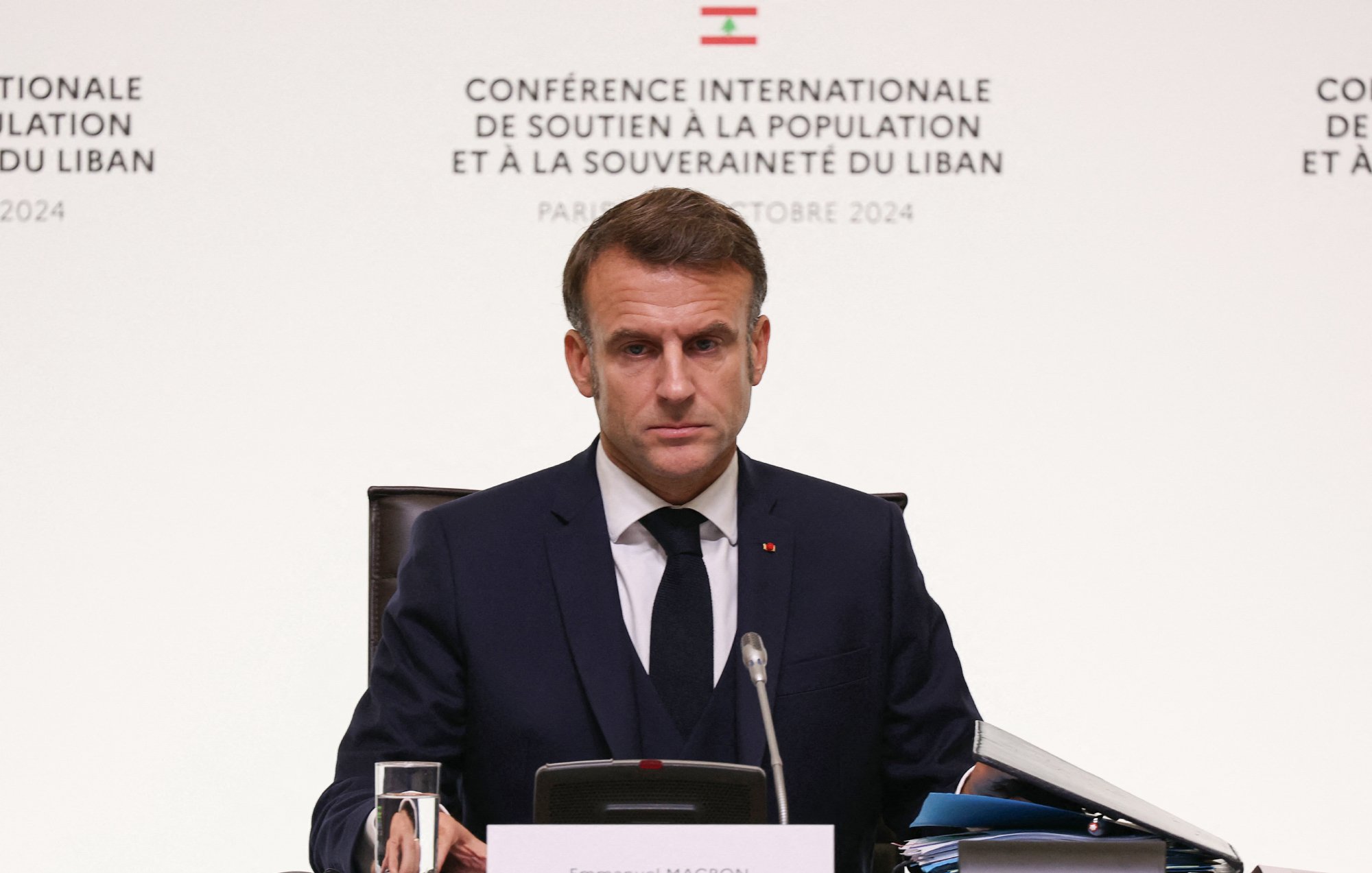 L'image montre un homme assis à une table lors d'une conférence. Il est habillé en costume sombre et porte une cravate. L'arrière-plan présente des inscriptions en français, indiquant qu'il s'agit d'une conférence internationale soutenant la population et la souveraineté du Liban, à Paris en octobre 2024. L'homme semble concentré et sérieux, regardant vers l'avant. Sur la table, on peut voir un verre d'eau et des documents.