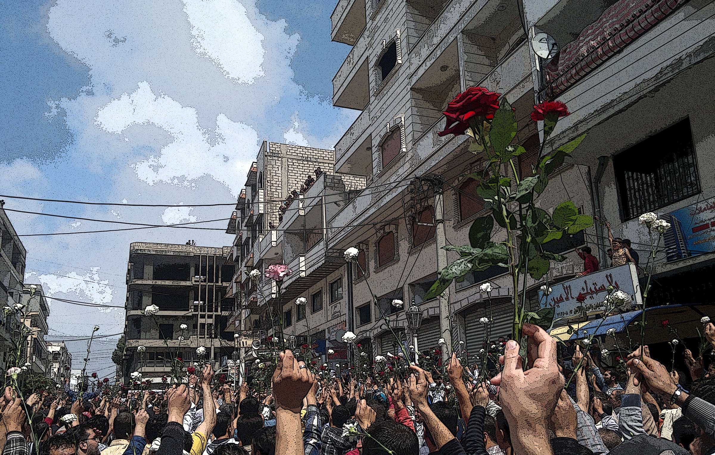 L'image montre une grande foule au milieu d'une rue, probablement lors d'une manifestation. Les gens semblent levés les bras, tenant des roses, ce qui suggère une ambiance pacifique ou festive. En arrière-plan, on aperçoit des bâtiments et un ciel partiellement nuageux. L'atmosphère semble être chargée d'émotions et d'unité entre les participants.