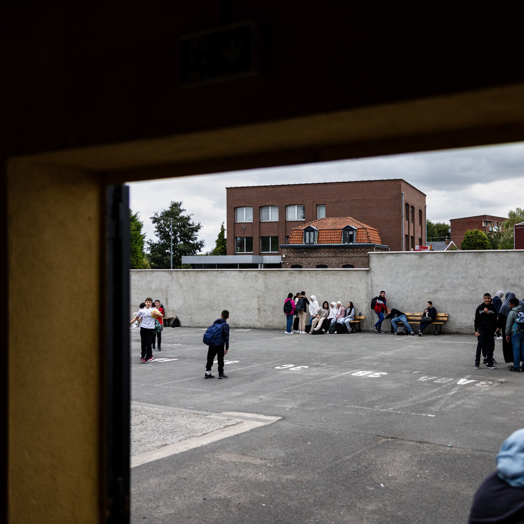 L'image montre une scène d'un espace extérieur, probablement une cour de récréation ou un terrain de sport. On peut voir plusieurs groupes de jeunes, répartis sur le sol, assis ou discutant ensemble. À l'arrière-plan, il y a des bâtiments avec des fenêtres et un toit de briques. Le ciel est nuageux, ce qui donne une ambiance un peu sombre à la scène. Les jeunes semblent engagés dans des conversations ou des activités sociales.