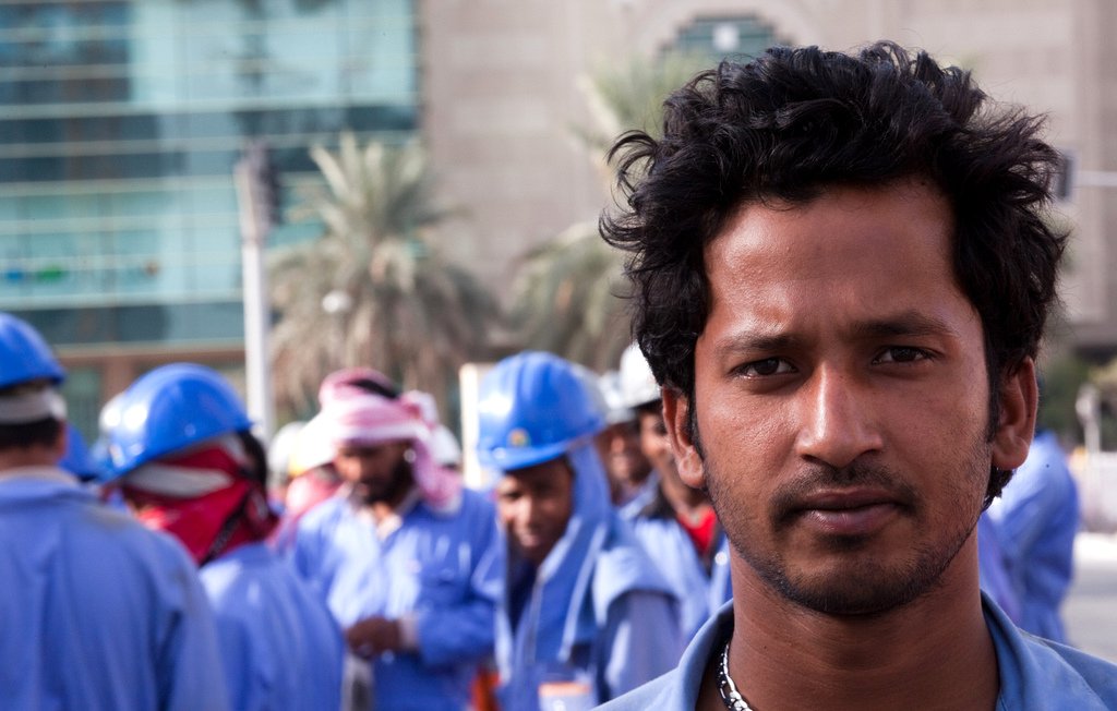 L'image montre un groupe de travailleurs, avec un homme au premier plan qui a les cheveux frisés et un regard intense. Il porte une tenue de travail bleue et une chaîne autour du cou. En arrière-plan, d'autres travailleurs en uniformes et casques bleus sont visibles, discutant entre eux. L'environnement semble urbain, avec des bâtiments modernes.