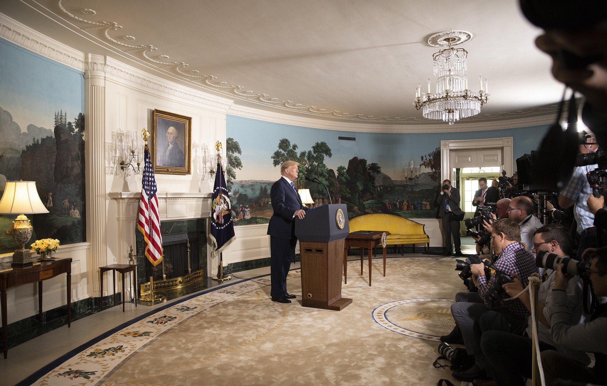 L'image montre un homme debout devant un podium, probablement lors d'une allocution publique. Il est situé dans une pièce décorée avec des murs peints de paysages, et des drapeaux américain et présidentiel sont visibles à côté de lui. Plusieurs journalistes et photographes sont présents, capturant l'événement. Le mobilier de la pièce semble ancien, et il y a un grand lustre au plafond.