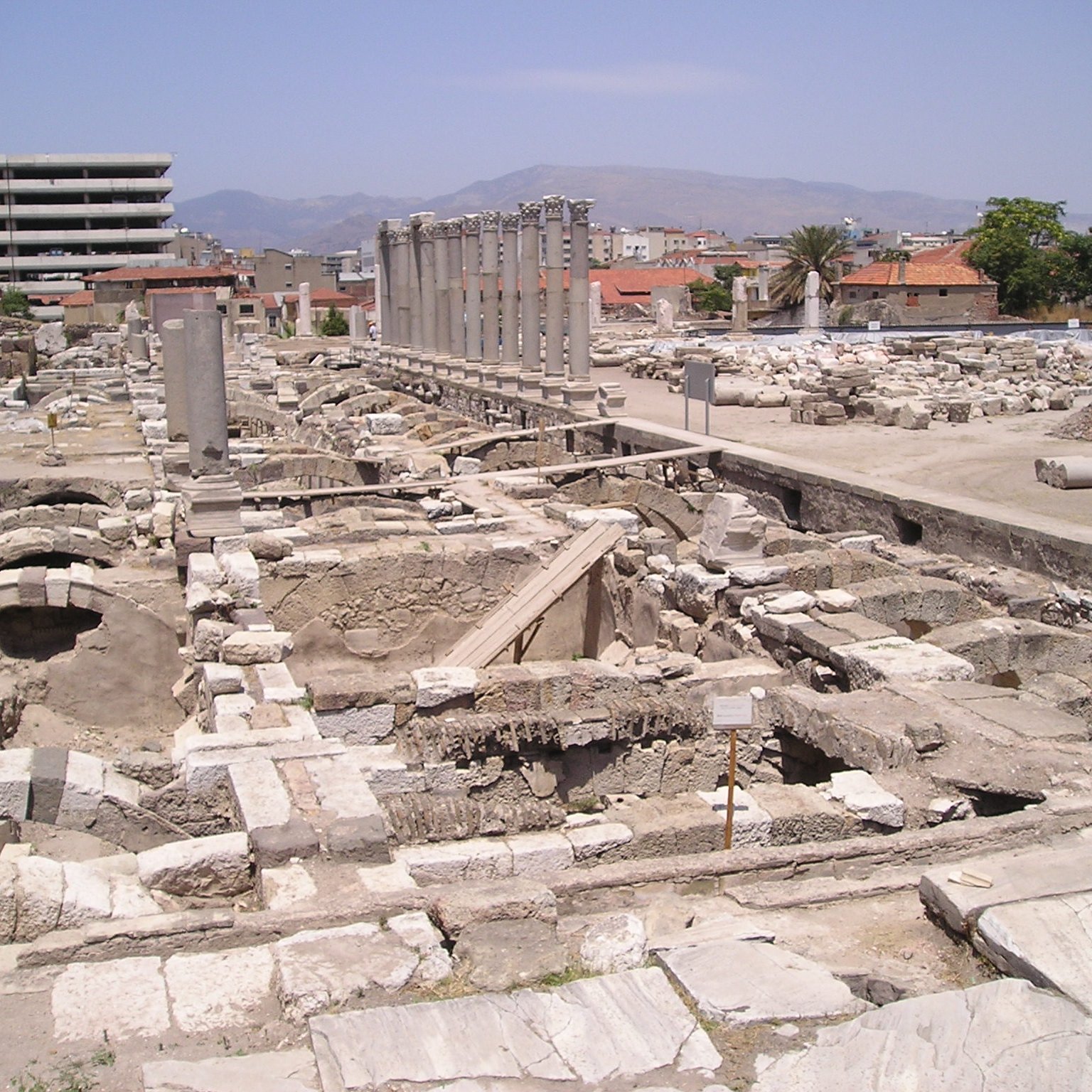 L'image montre un site archéologique avec des ruines et des structures anciennes. On aperçoit des colonnes dressées, probablement d'un bâtiment antique, ainsi que des fondations de murs en pierre. En arrière-plan, on peut distinguer des bâtiments modernes, contrastant avec les vestiges historiques. Le ciel est clair et le paysage est légèrement vallonné au loin. L'endroit semble être une zone de fouilles, avec des éléments qui témoignent d'une ancienne civilisation.