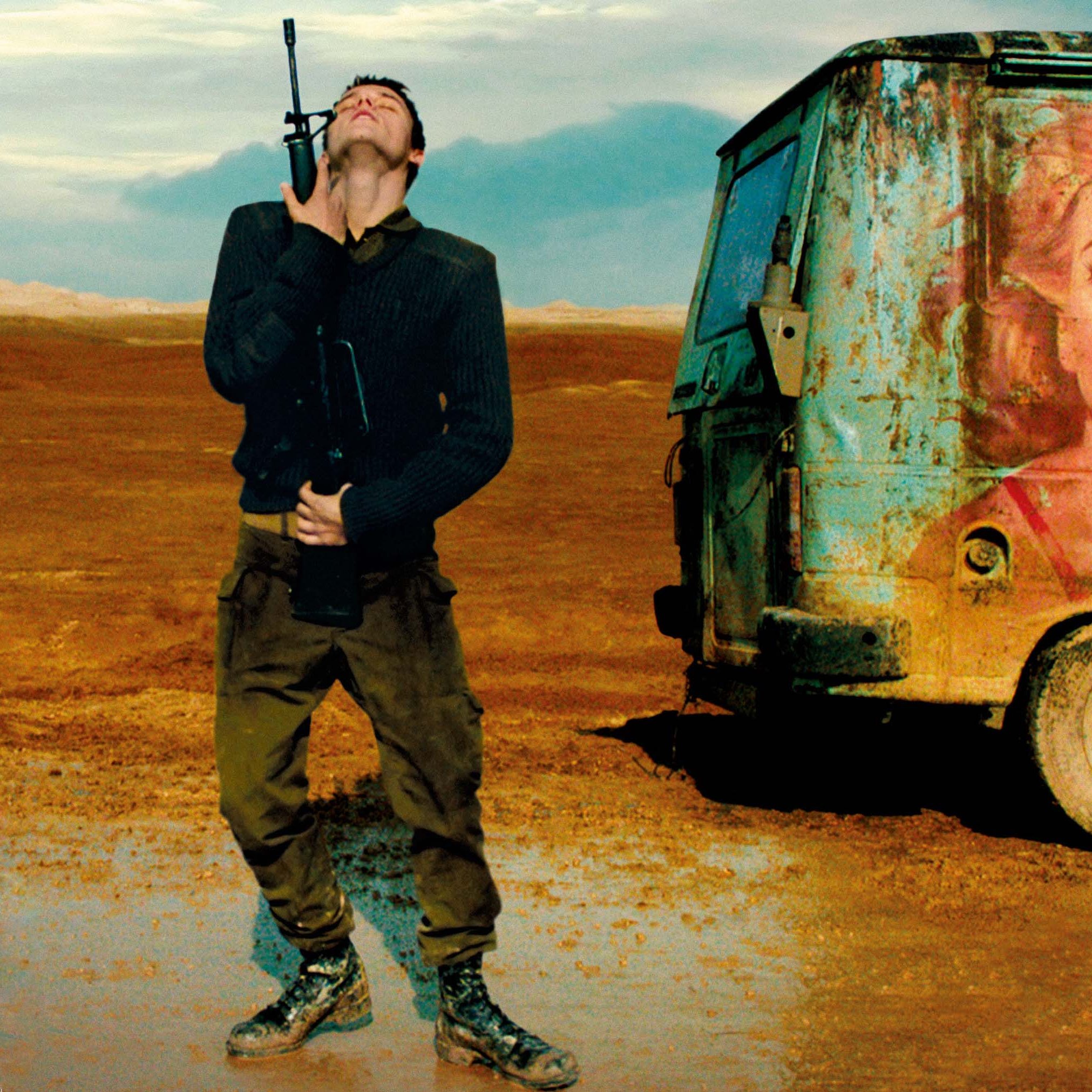 L'image montre un homme debout dans un paysage désertique, avec une expression qui semble mélancolique ou contemplative. Il porte une arme à feu à la main et est habillé de manière militaire. À côté de lui se trouve un vieux camion, sur lequel est peint un portrait coloré d'une femme souriante tenant une glace. Le contraste entre l'aspect rustique du véhicule et l'image joyeuse de la femme crée une atmosphère intrigante, mêlant des éléments de décor et d'émotion. Le ciel, quant à lui, est nuageux, ajoutant une dimension dramatique à la scène.