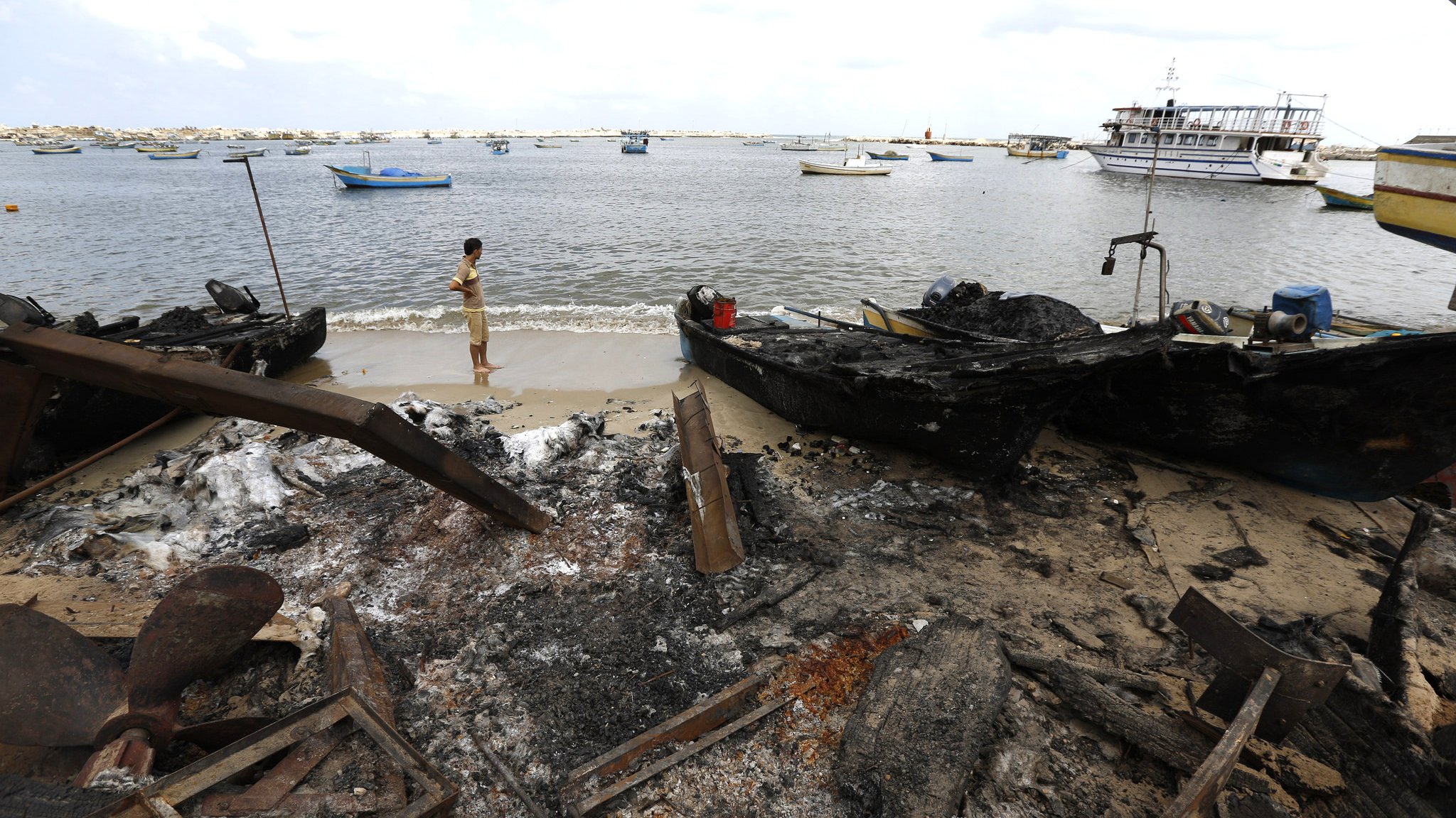 الصورة تُظهر مشهداً لواجهة مائية حيث يبدو أن هناك قوارب محترقة على الشاطئ. فيforeground، يظهر شخص واقفًا ينظر إلى المياه. تُظهر الخلفية قوارب أخرى مائية ومشهدًا طبيعيًا مع سماء ملبدة بالغيوم. يبدو أن الوضع يشير إلى أثر الدمار أو الحريق على البيئة البحرية.