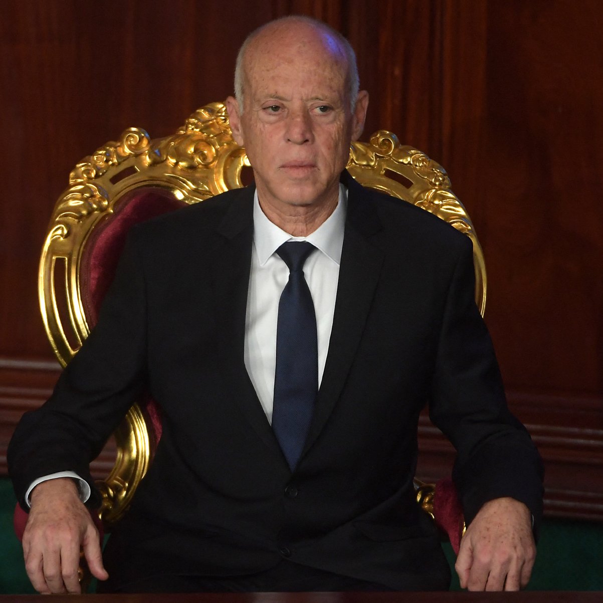 L'image montre un homme assis sur une grande chaise ornée, avec un décor en bois sombre en arrière-plan. Il porte un costume noir et une cravate sombre, affichant une expression sérieuse. Son attitude et son cadre suggèrent une ambiance officielle ou formelle.