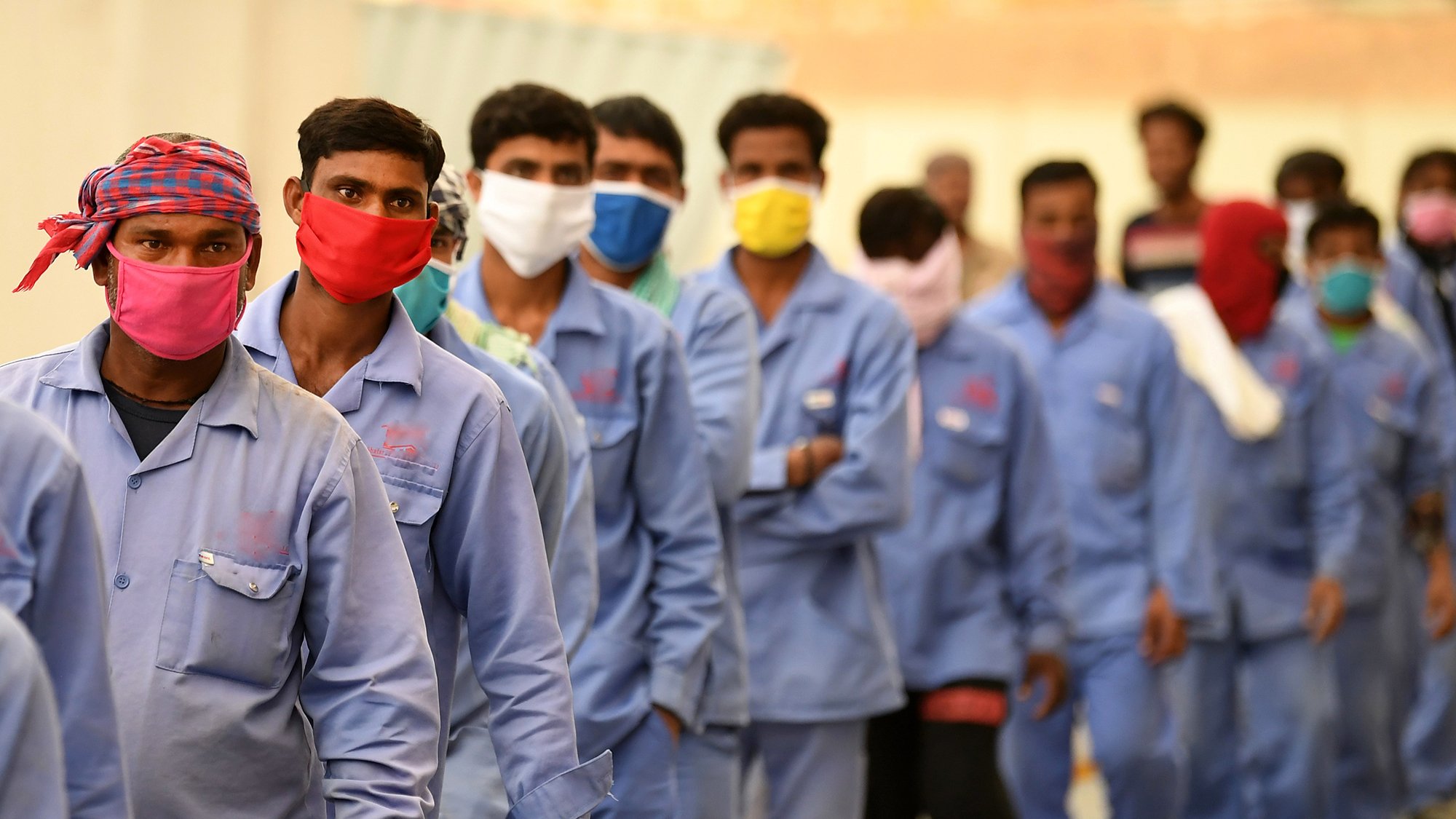 En la imagen se puede observar a un grupo de hombres alineados, todos ellos usando mascarillas de diferentes colores. Visten uniformes azules y parecen estar en un entorno al aire libre. La fila de personas avanza hacia algún destino, y el ambiente sugiere que podría ser una situación laboral o de espera. Algunos de los hombres llevan pañoletas en la cabeza, lo que indica posibles medidas de protección o identificación.