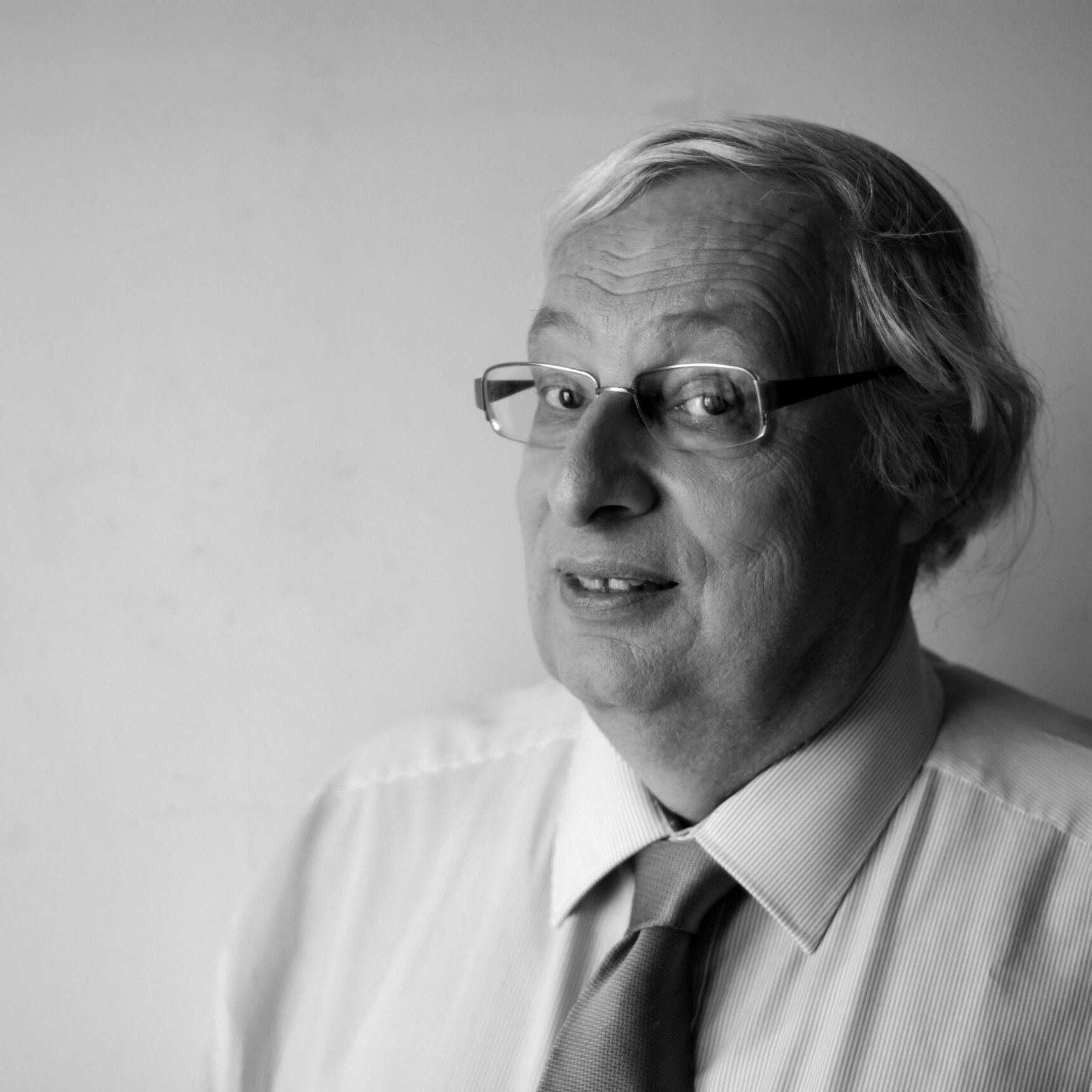 L'image montre un homme d'âge moyen avec des cheveux gris et des lunettes. Il porte une chemise claire avec une cravate. Il regarde légèrement sur le côté avec un sourire, le fond étant neutre et clair. L'image est en noir et blanc, ce qui lui donne un aspect classique et intemporel.