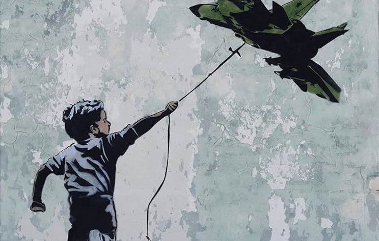 L'immagine rappresenta un bambino che corre e tiene in mano un filo da cui pende un aereo da guerra in miniatura. Il bambino sembra gioioso, mentre lo sfondo è costituito da un muro scrostato con tonalità chiare e imperfezioni visibili. L'aereo, di colore verde militare, contrasta con l'innocenza del bambino e il suo gesto, creando una riflessione evocativa sul tema della guerra e della gioventù.