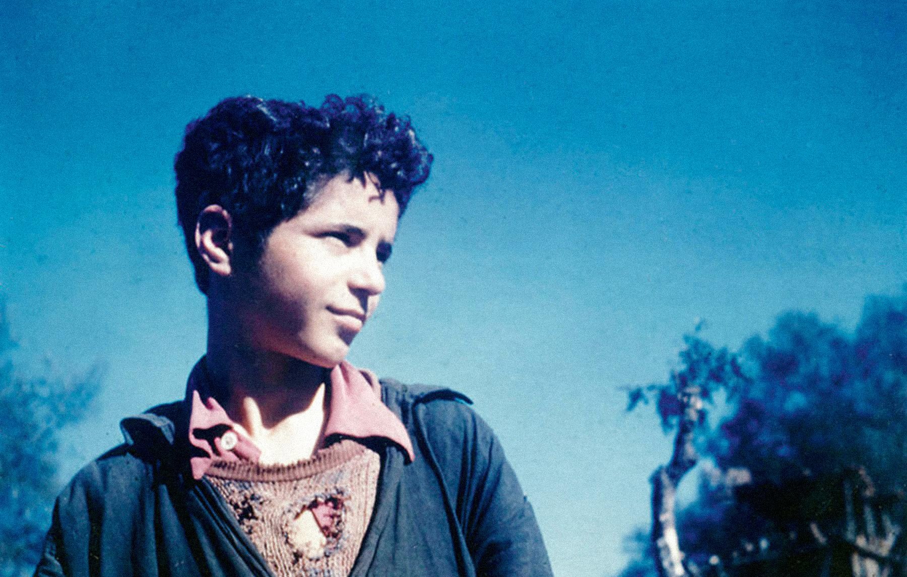 L'image présente un jeune garçon au visage expressif, avec des cheveux frisés et un regard tourné légèrement sur le côté. Il porte un vêtement sombre avec un col de chemise visible en dessous. L'arrière-plan est flou, aux teintes de bleu, ce qui crée une ambiance douce et rêveuse. Le garçon semble pensif ou contemplatif, se détachant bien dans cette atmosphère colorée.