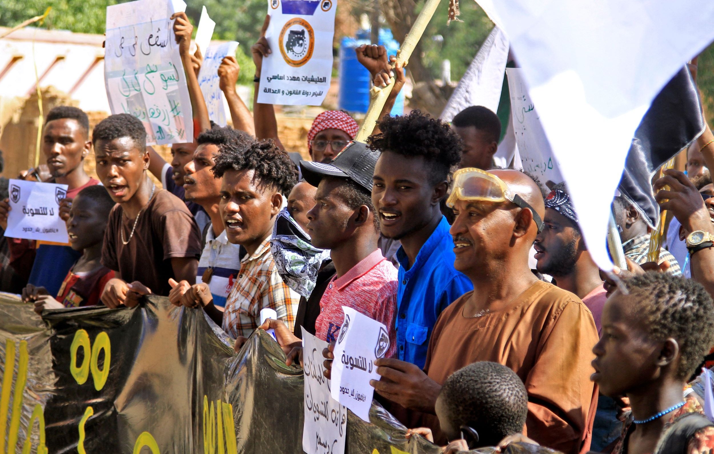 L'immagine ritrae una folla di manifestanti che tiene in mano cartelli e striscioni. I partecipanti sembrano coinvolti ed esprimono entusiasmo, alcuni sorridono mentre altri alzano le mani. Sono visibili diverse espressioni facciali e abbigliamento vario, con colori e stili che riflettono un'atmosfera di protesta. Sullo sfondo si intravede un ambiente urbano, con edifici e vegetazione. L'atteggiamento dei manifestanti suggerisce che stanno lottando per una causa importante.