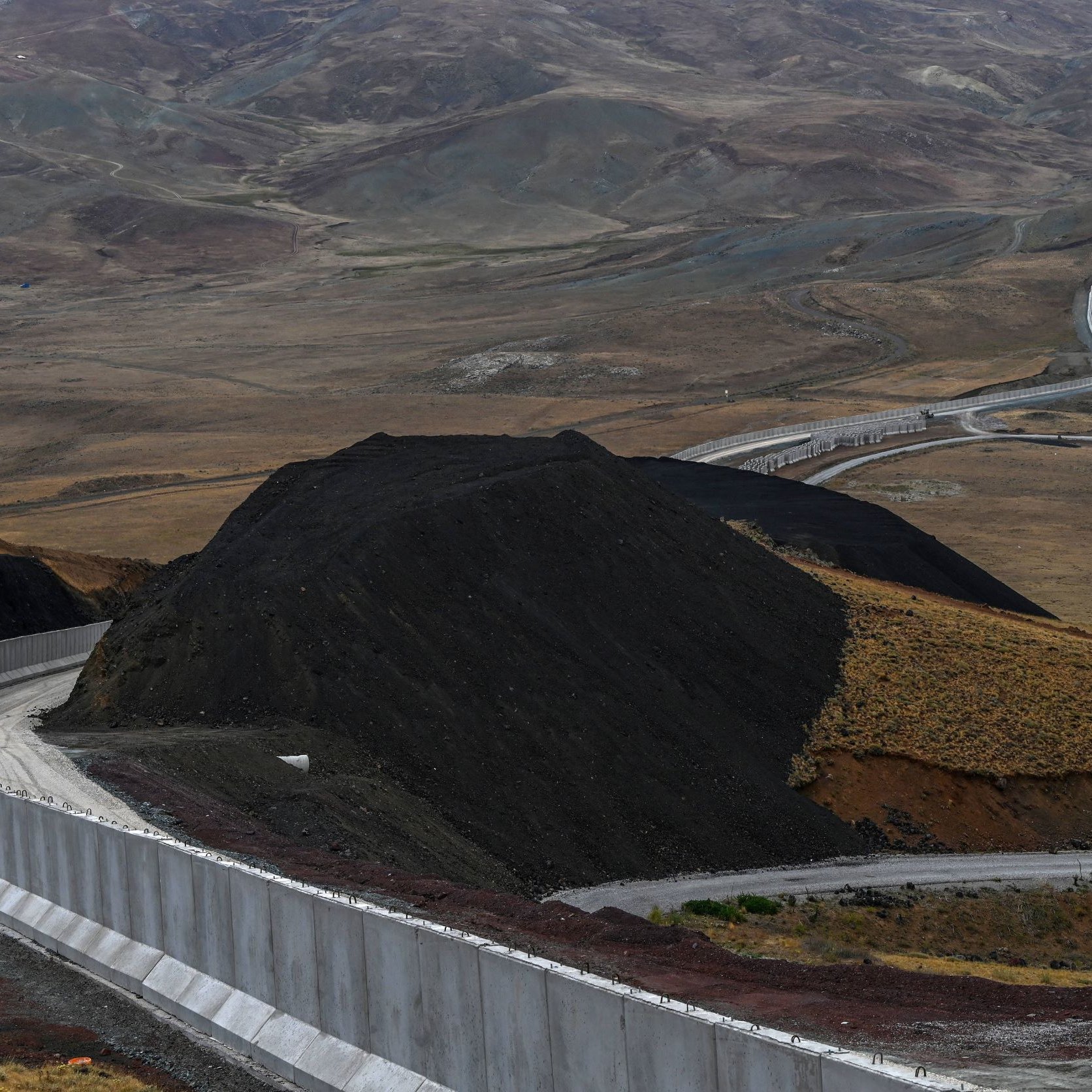 L'immagine mostra un paesaggio montano caratterizzato da una strada che si snoda attraverso un terreno collinare. Sullo sfondo, si possono vedere colline e valli, mentre in primo piano c'è una parete di cemento che segue il contorno della strada. Il terreno appare secco e brullo, con tonalità di marrone e verde. La scena comunica un senso di isolamento e vastità, tipico di un ambiente montano.