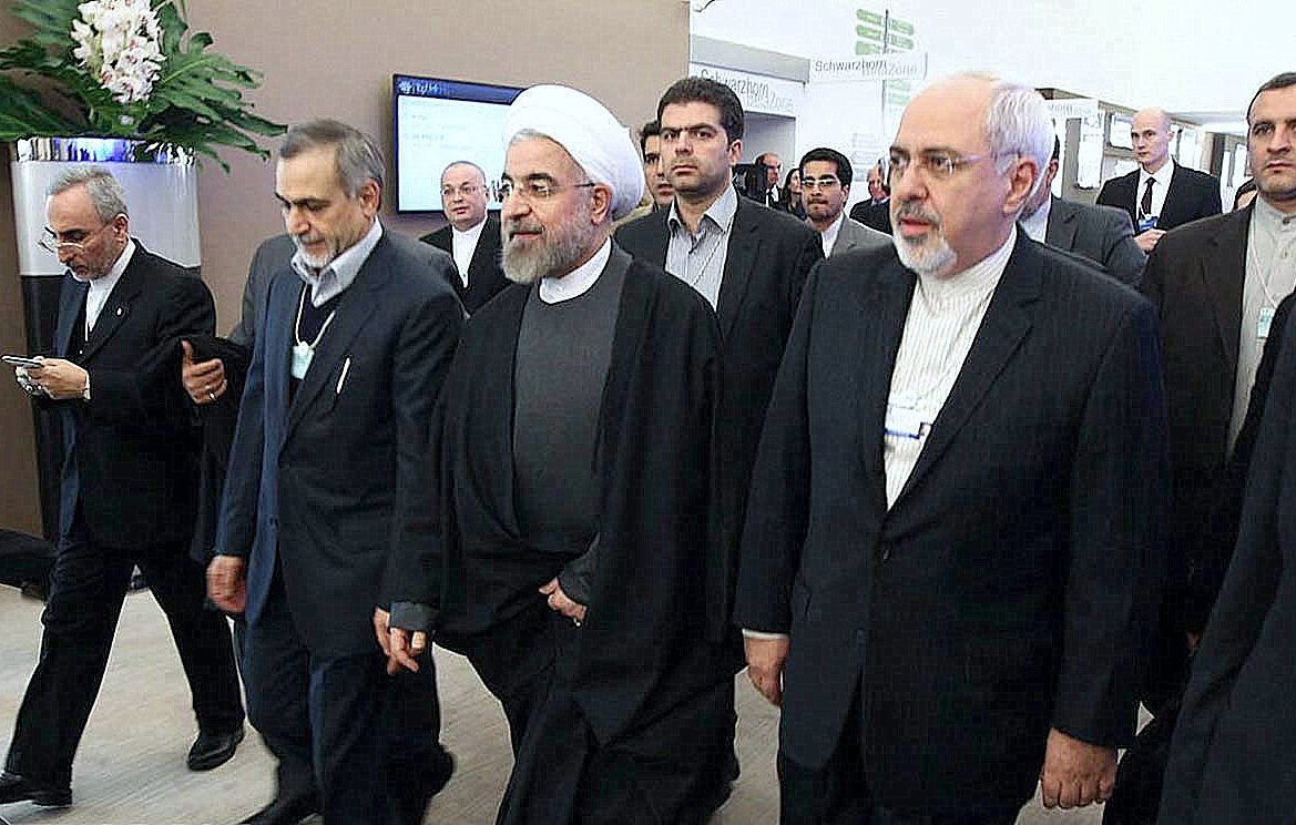 L'image montre un groupe d'hommes en costume formel, marchant ensemble dans un environnement apparemment professionnel. Certains d'entre eux portent des vêtements traditionnels, comme des chapeaux ou des manteaux, tandis que d'autres ont des costumes occidentaux. L'atmosphère semble être celle d'un événement officiel ou d'une conférence, avec des personnes à l'arrière-plan qui observent ou participent également. Leur expression indique une certaine concentration et un engagement dans la situation.