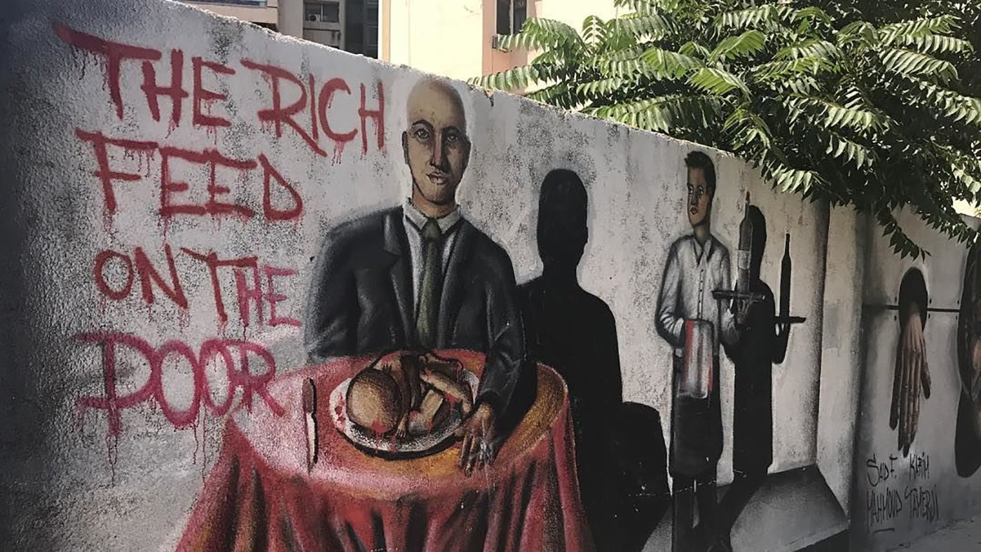 L'image montre une fresque murale colorée sur un mur. Au premier plan, un homme en costume est assis à une table couverte d'une nappe rouge, avec un plat à manger devant lui. À côté, on peut voir une ombre d'un serveur, suggérant un contexte de service alimentaire. Sur le mur, il est écrit "THE RICH FEED ON THE POOR", ce qui évoque un message critique sur les inégalités sociales. Des éléments de l'arrière-plan, comme des bâtiments et de la verdure, ajoutent à l'environnement urbain.