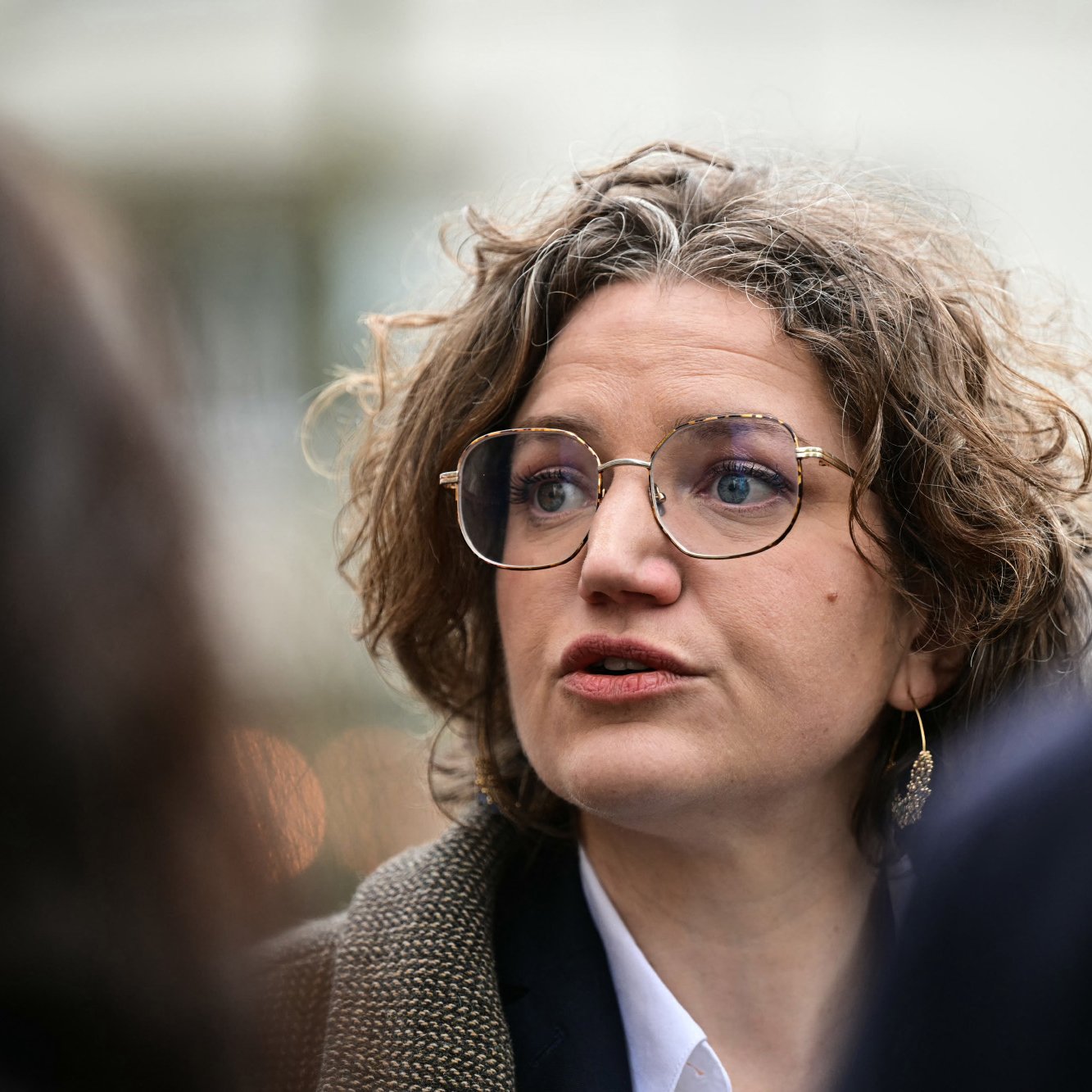 L'image montre une femme avec des cheveux bouclés et des lunettes, en train de parler. Elle semble engagée dans une conversation, peut-être lors d'un événement public. Son expression faciale est sérieuse, et elle porte un vêtement avec un col blanc et une veste. L'arrière-plan paraît flou, suggérant qu'elle se trouve au milieu d'un groupe ou d'une foule.