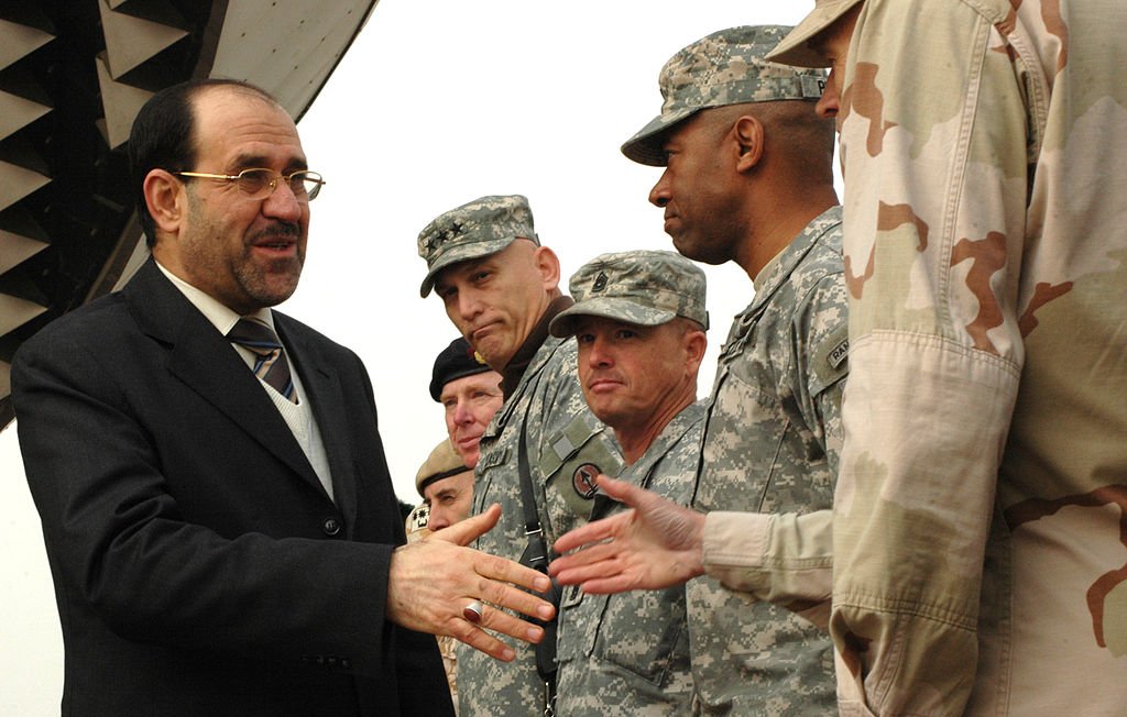 L'image montre un homme en costume qui serre la main de plusieurs militaires. L'homme semble être en discussion amicale avec eux. Les soldats portent des uniformes militaires, et l'environnement semble être une zone aéroportuaire, avec un avion visible en arrière-plan. Les expressions des militaires sont sérieuses, tandis que l'homme en costume sourit. Ce moment pourrait symboliser une collaboration ou une rencontre officielle.