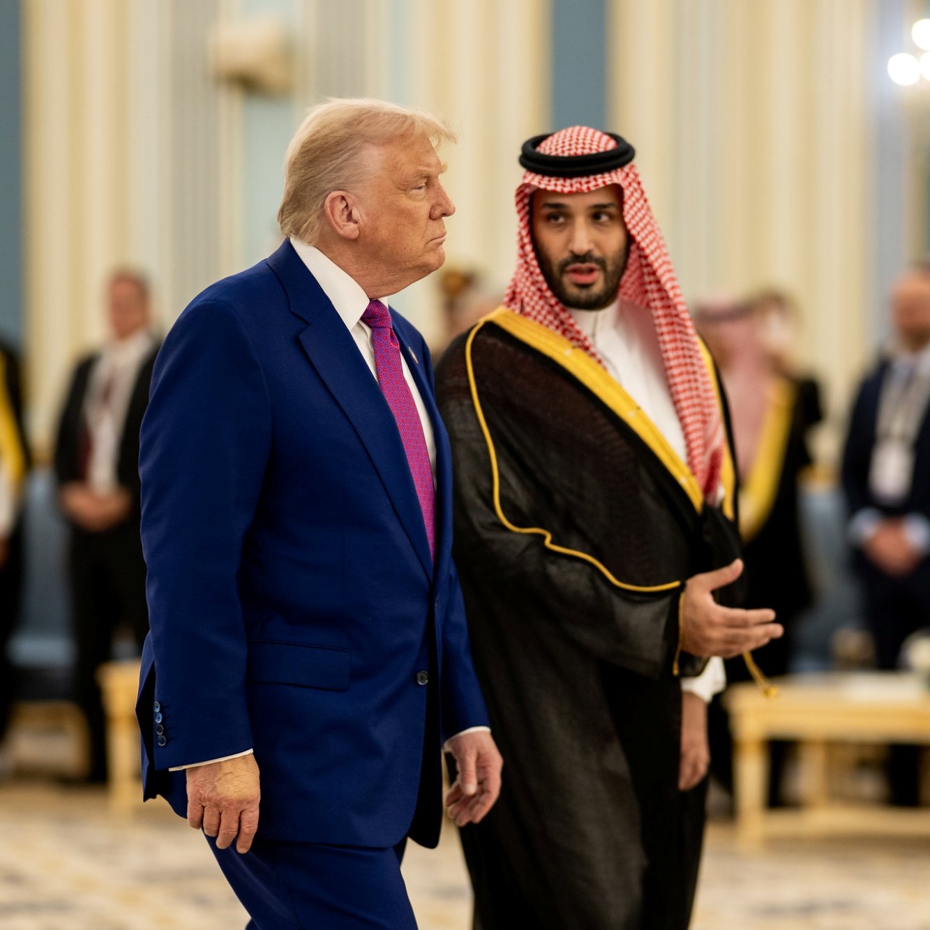 دو مرد در لباس‌های رسمی مختلف در یک محیط مجلل با نور ملایم و حضور افرادی دیگر.