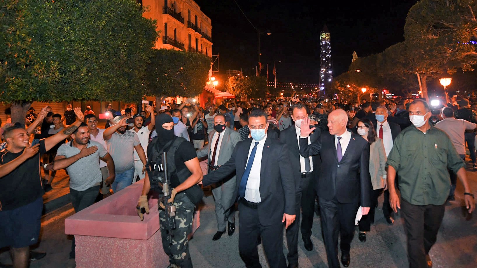 تظهر الصورة مجموعة من الأشخاص في شوارع مدينة مضاءة بأضواء ليلية. يتواجد في المقدمة رجل يرتدي بدلة مع أشخاص آخرين، من بينهم رجل ملثم يحمل سلاحًا. يبدو أن هناك تجمعًا من الحشود في الخلفية، حيث يتفاعل بعضهم مع الموقف. الأجواء توحي بوجود حدث أو مظاهرة، ومشاعر الحماس والانفعال واضحة في تعابير الحضور.