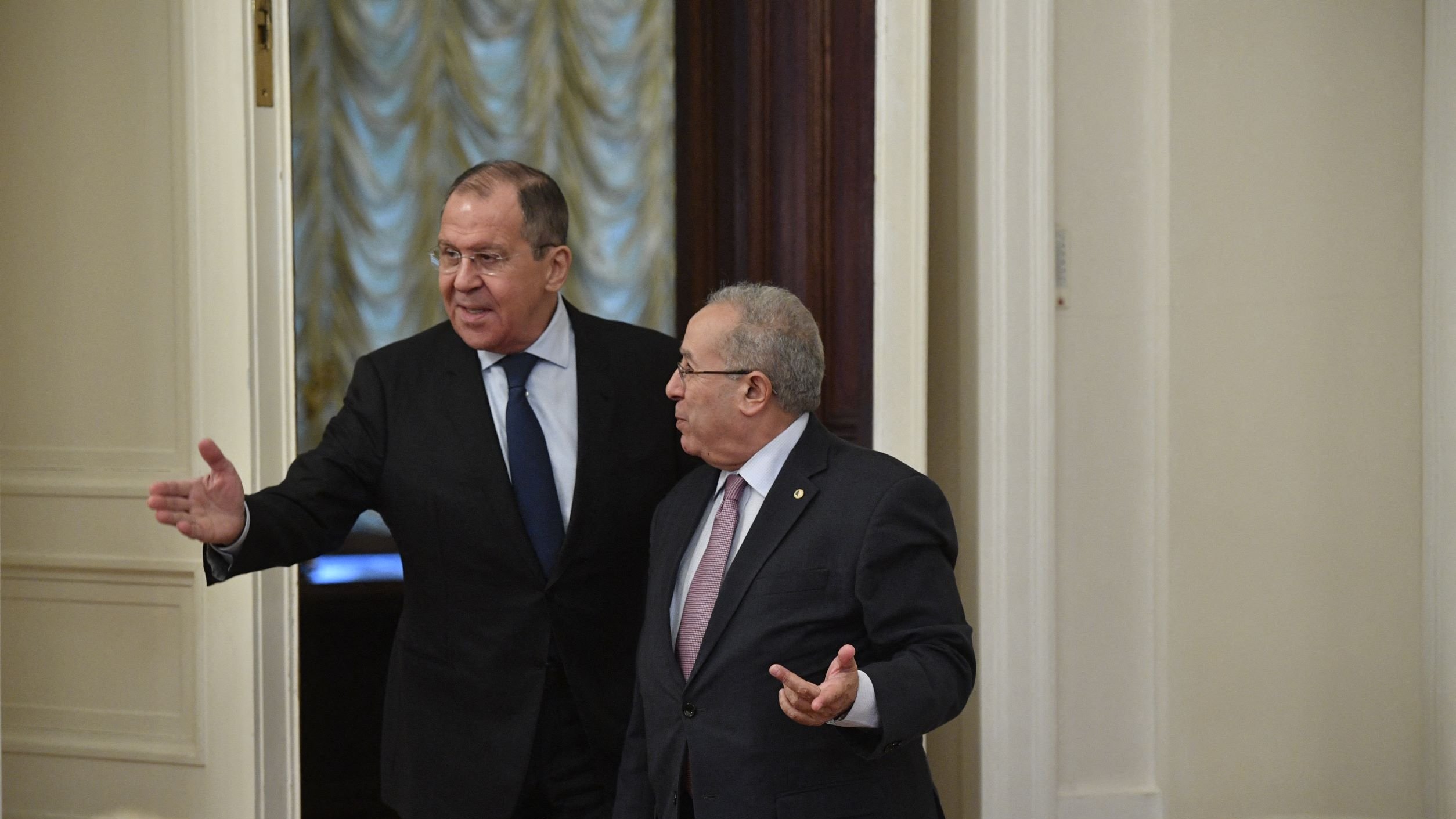 این تصویر دو مرد را نشان می‌دهد که در یک فضای داخلی ملاقات می‌کنند. یکی از آنها لباس رسمی با کت و شلوار به تن دارد و به سمت دوربین اشاره می‌کند. دیگری نیز در حالی که لباسی رسمی به تن دارد، به طرف مقابل نگاه می‌کند و دستش را به نشانه صحبت دراز کرده است. محیط اطراف شامل دیوارهایی با طراحی کلاسیک و پرده‌هایی به رنگ ملایم است. به نظر می‌رسد این ملاقات در یک مکان رسمی انجام می‌شود.