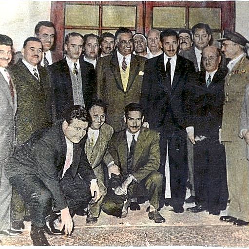 L'image montre un groupe de plusieurs hommes rassemblés dans une pièce. Ils portent des vêtements variés, allant de costumes formels à des tenues traditionnelles. Certains se tiennent debout tandis que d'autres sont accroupis ou assis. Le décor semble être d'une certaine importance, potentiellement une cérémonie ou un événement officiel. Les expressions faciales des personnes varient, allant de sourires à des regards sérieux, ce qui suggère un mélange de convivialité et de gravité. Les détails sur les personnalités présentes et le contexte de l'image ne sont pas précisés.