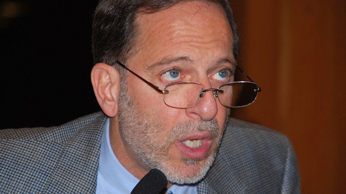 Je ne sais pas qui est cette personne. Cependant, je peux dire que l'image semble montrer un homme portant des lunettes, avec une barbe grisonnante et des cheveux également grisonnants. Son expression semble sérieuse et il est peut-être en train de parler ou de faire une présentation. Son veston est en tissu à motifs.
